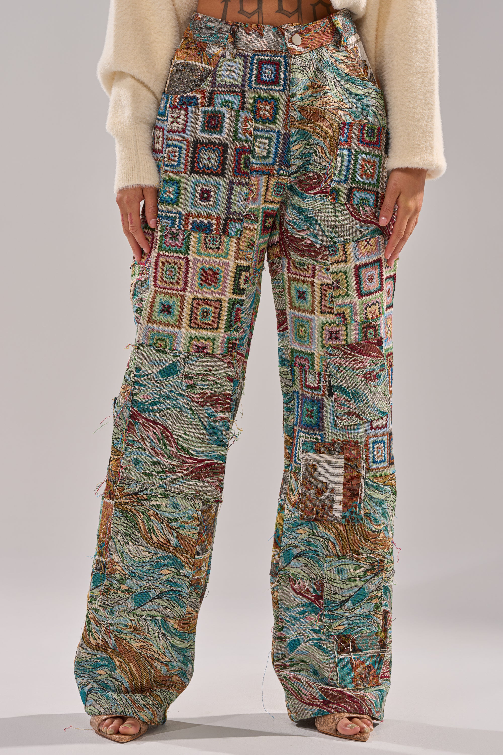 A person wears the WILD PATCHWORK TROUSER—high-waisted, wide-leg pants with colorful squares, swirls, and abstract patterns—paired with a cream fuzzy sweater; photo cropped at shoulders and feet.