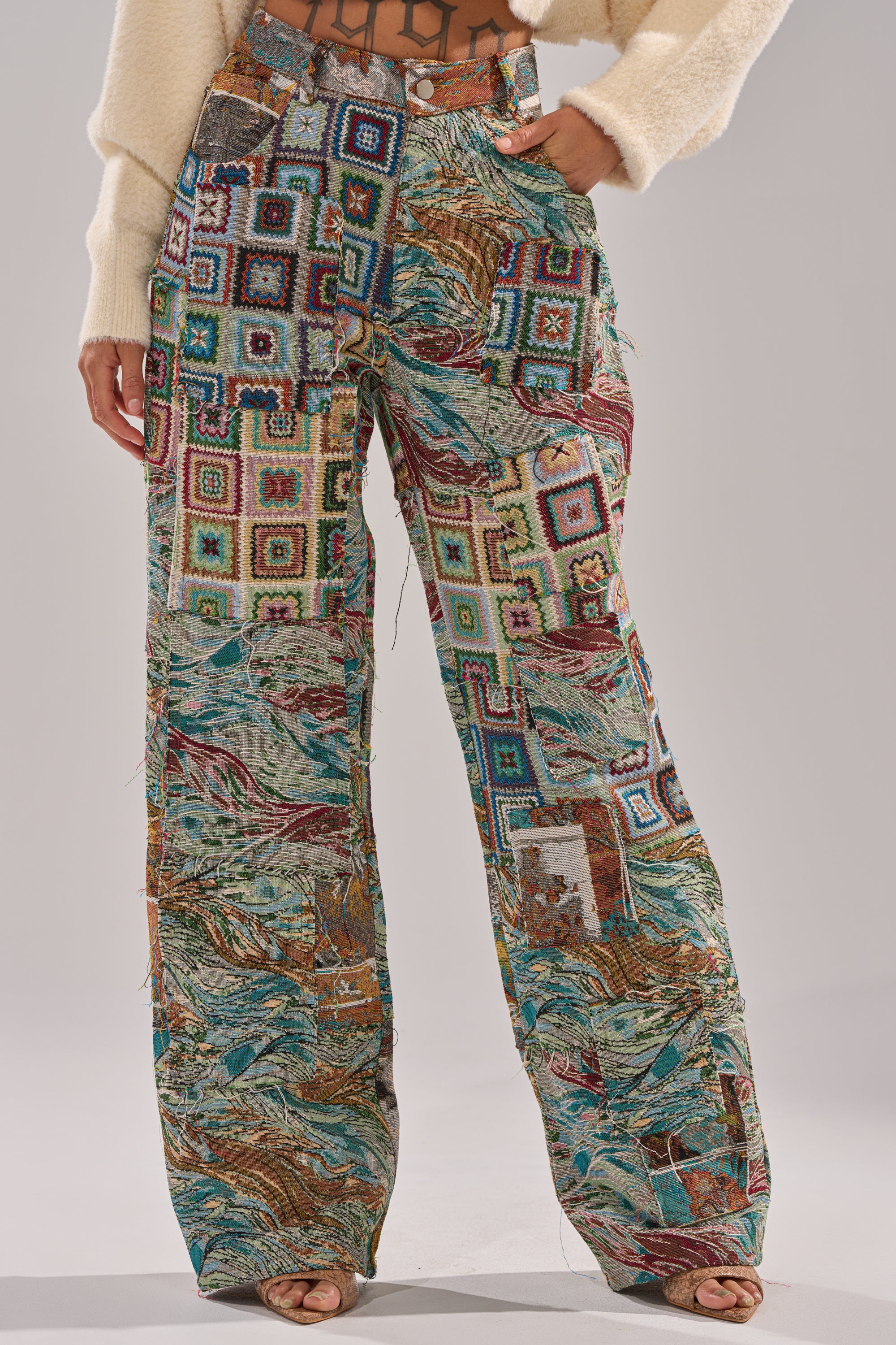 A person wears the WILD PATCHWORK TROUSER, a vintage-inspired high-waisted wide-leg pant with colorful crochet squares and abstract patterns, paired with a cream sweater. Only their torso and legs are visible.