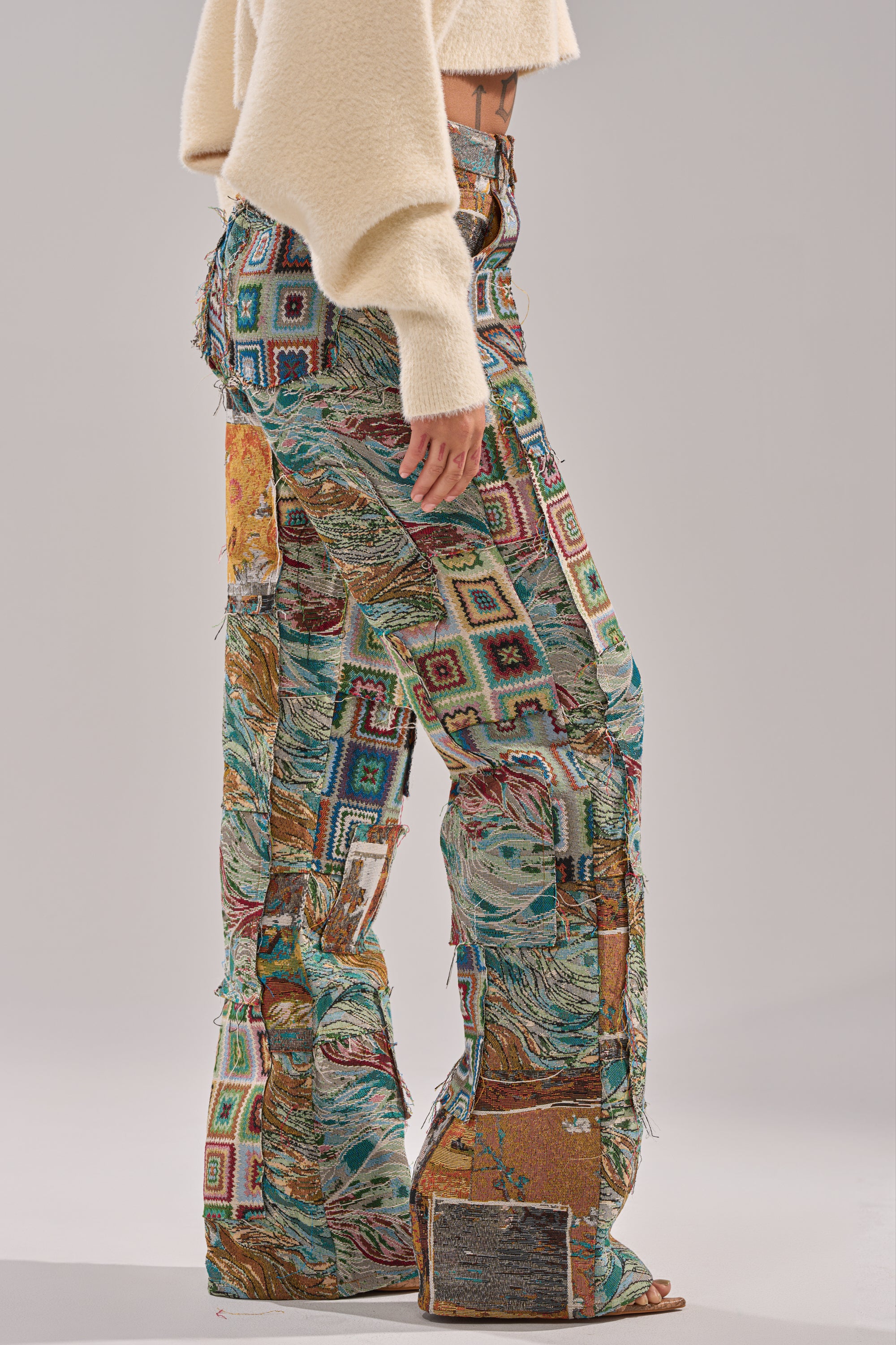 A person stands sideways, showing only their body from shoulders down, wearing WILD PATCHWORK TROUSER with a wide-leg silhouette, tapestry-style jacquard fabric, and a cream cropped sweater.