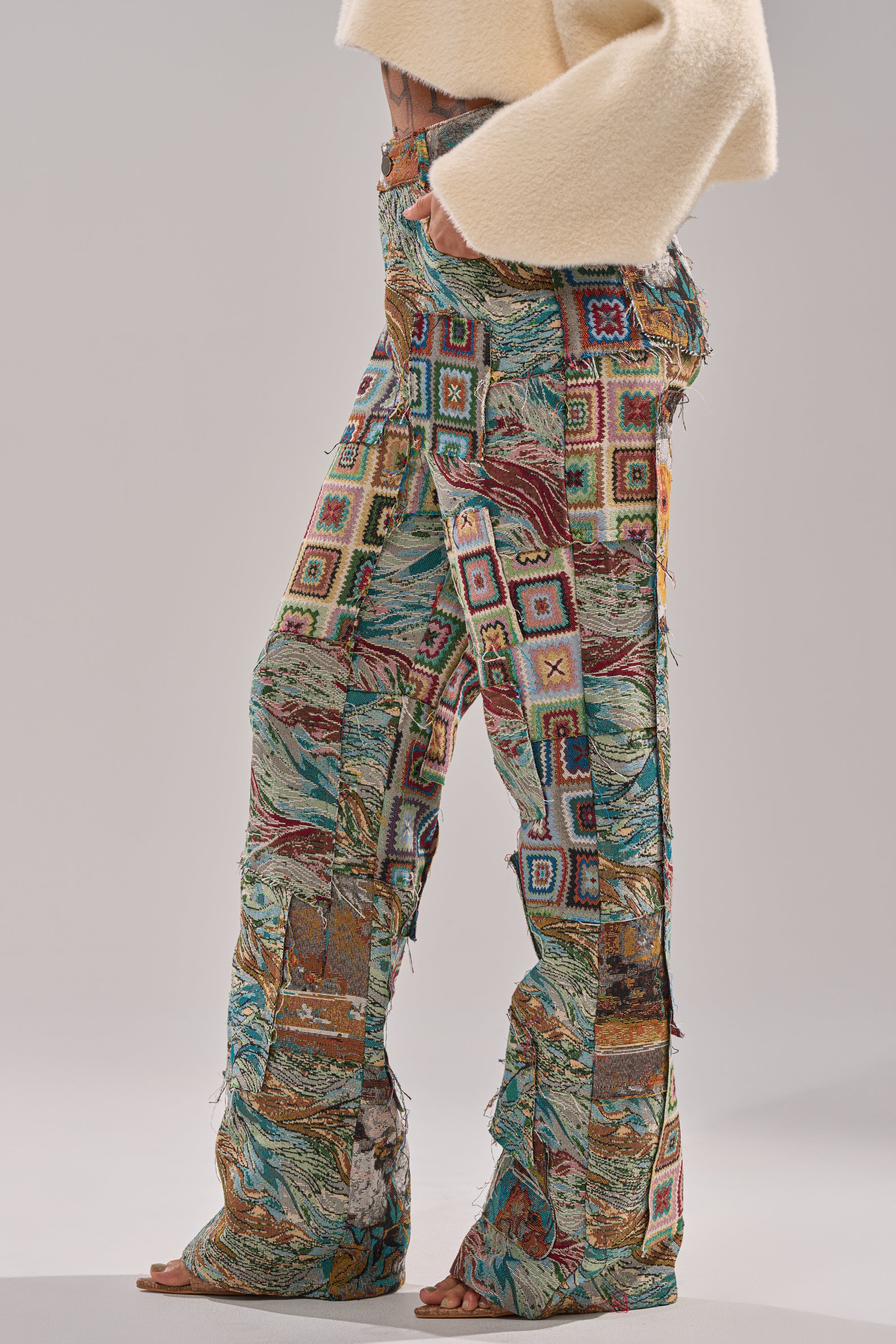 A person models the WILD PATCHWORK TROUSER, featuring a wide-leg silhouette and vibrant tapestry jacquard patterns, paired with a cream fuzzy cropped sweater. The image spotlights the lower body and legs.