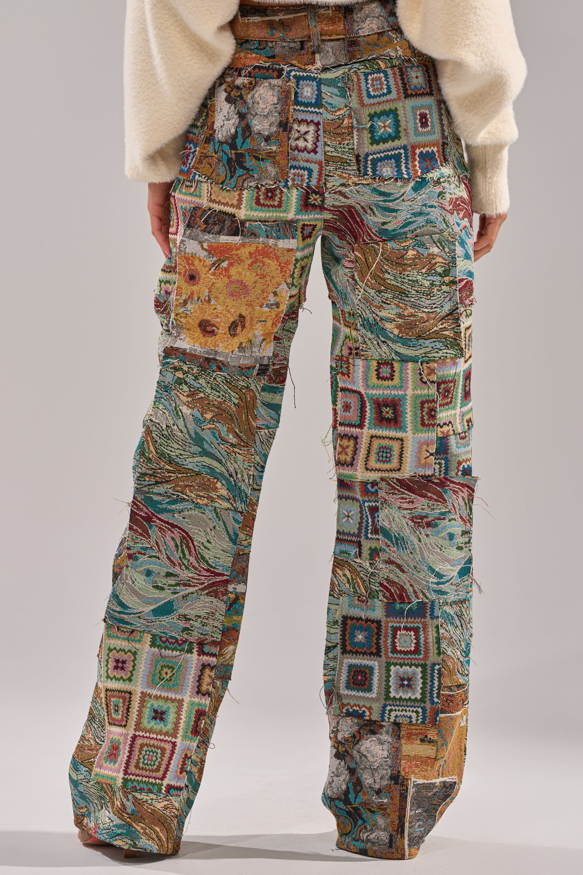 A person models the WILD PATCHWORK TROUSER, showcasing its vibrant green, blue, red, yellow, and beige squares with abstract patterns. Their cream sweater peeks out as the wide-leg silhouette is displayed from waist to feet.