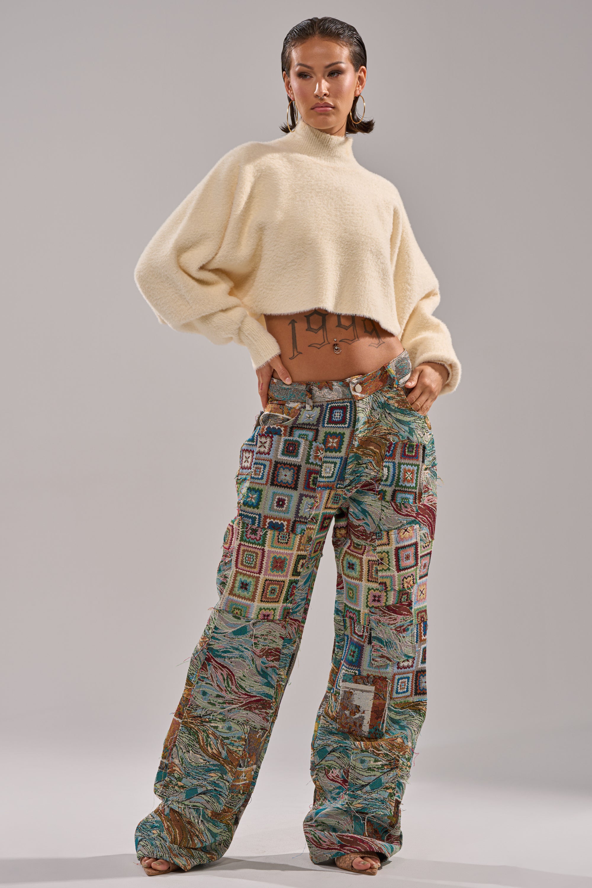 A person stands confidently in a cream, long-sleeve cropped sweater and WILD PATCHWORK TROUSER featuring a wide-leg silhouette. Slicked-back hair and visible torso tattoos add to their bold look.