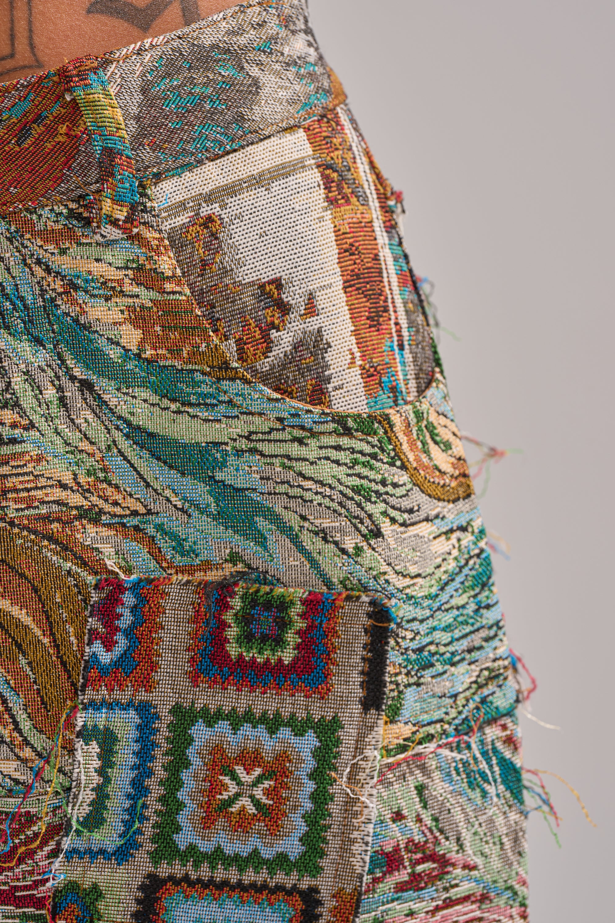 Close-up of a colorful, textured pocket on the WILD PATCHWORK TROUSER, showcasing intricate jacquard tapestry patterns with frayed edges. A phone in a matching patterned case peeks out from the pocket.