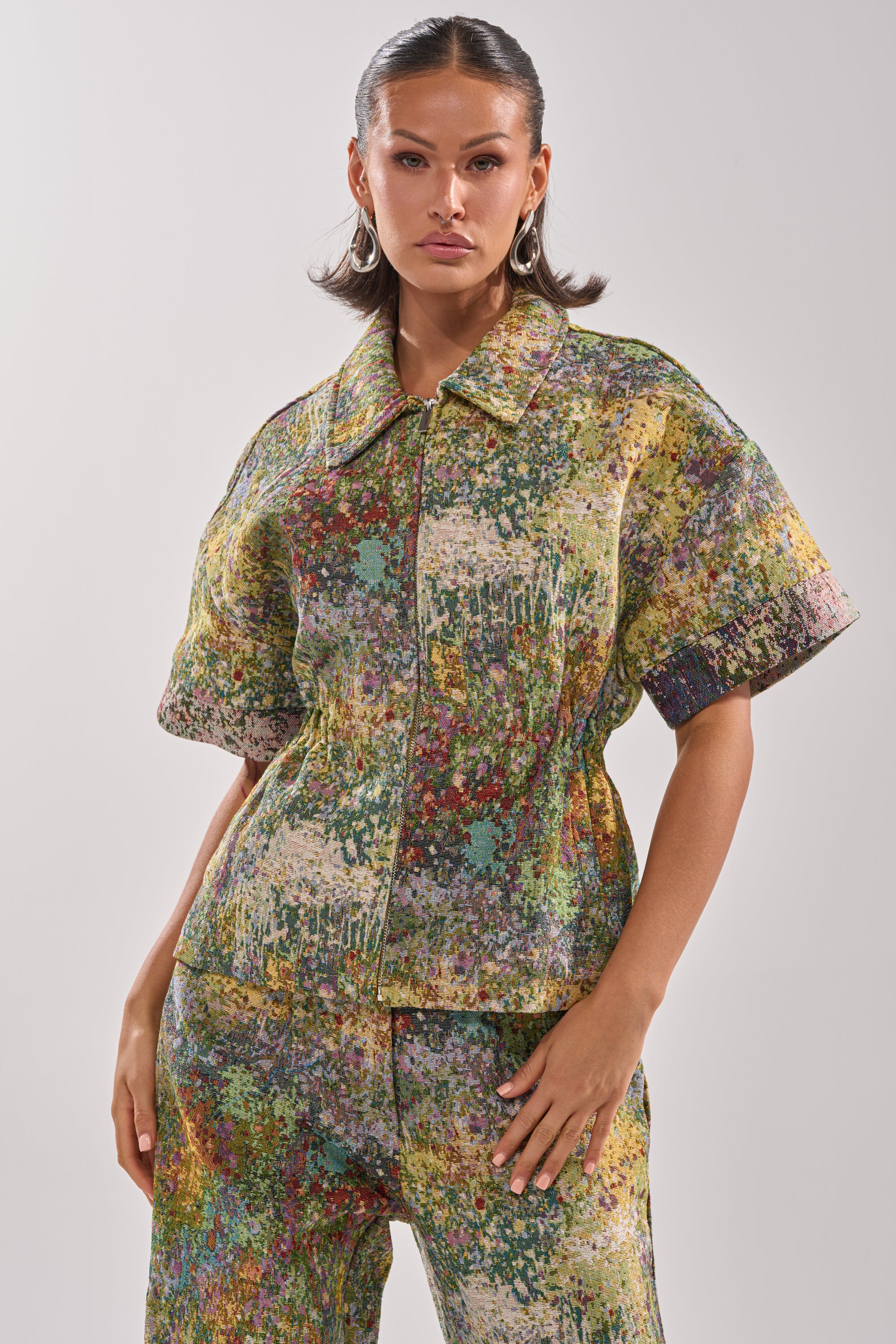 A woman wears the DEEP IN THE SEA BROCADE BLOUSE, featuring colorful, textured abstract patterns. She stands confidently with one hand by her side against a plain background.