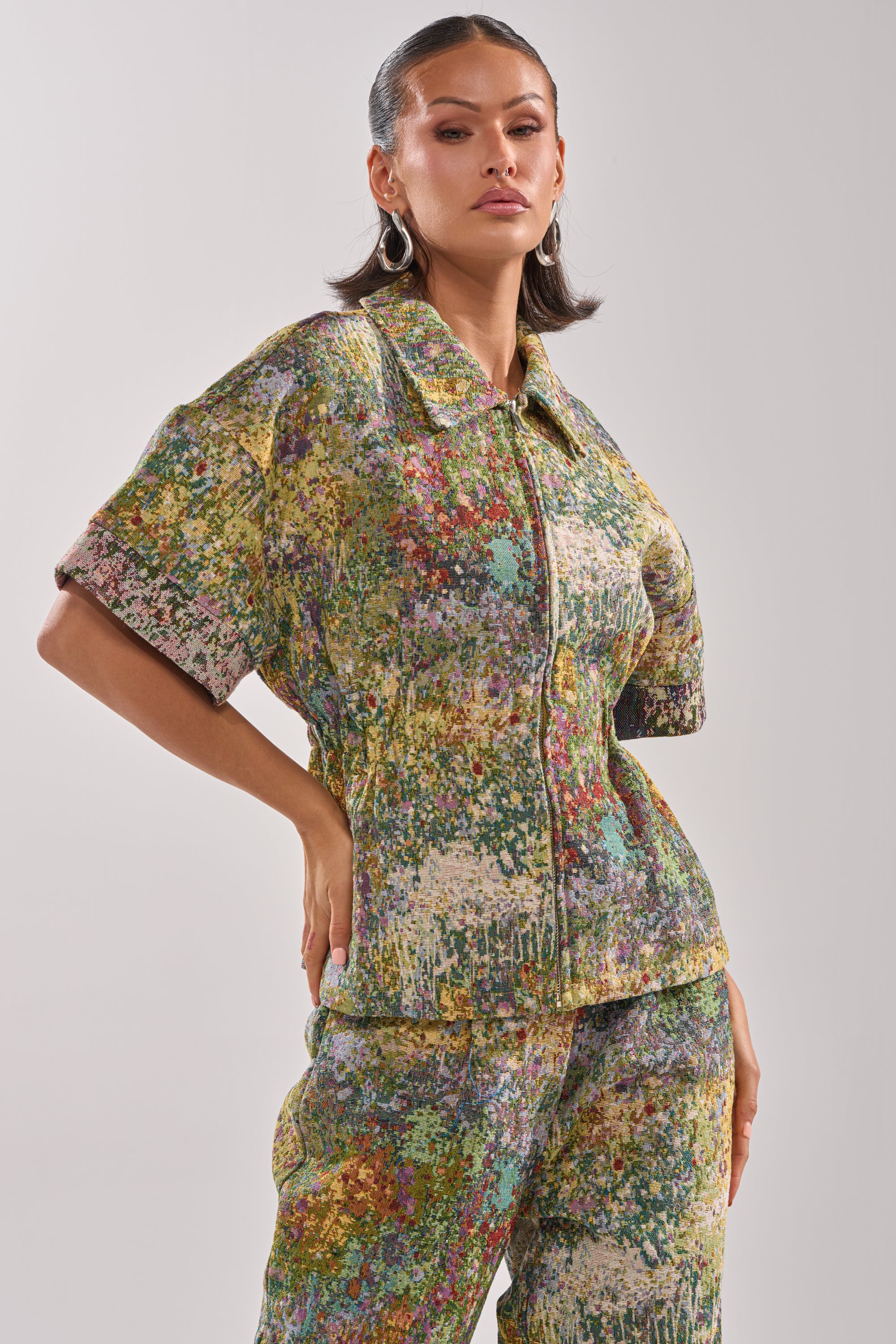 A woman wears the DEEP IN THE SEA BROCADE BLOUSE, a vibrant abstract floral top paired with matching pants and a short-sleeve zip-up jacket. She completes the look with sleek hair, hoop earrings, and a neutral expression.