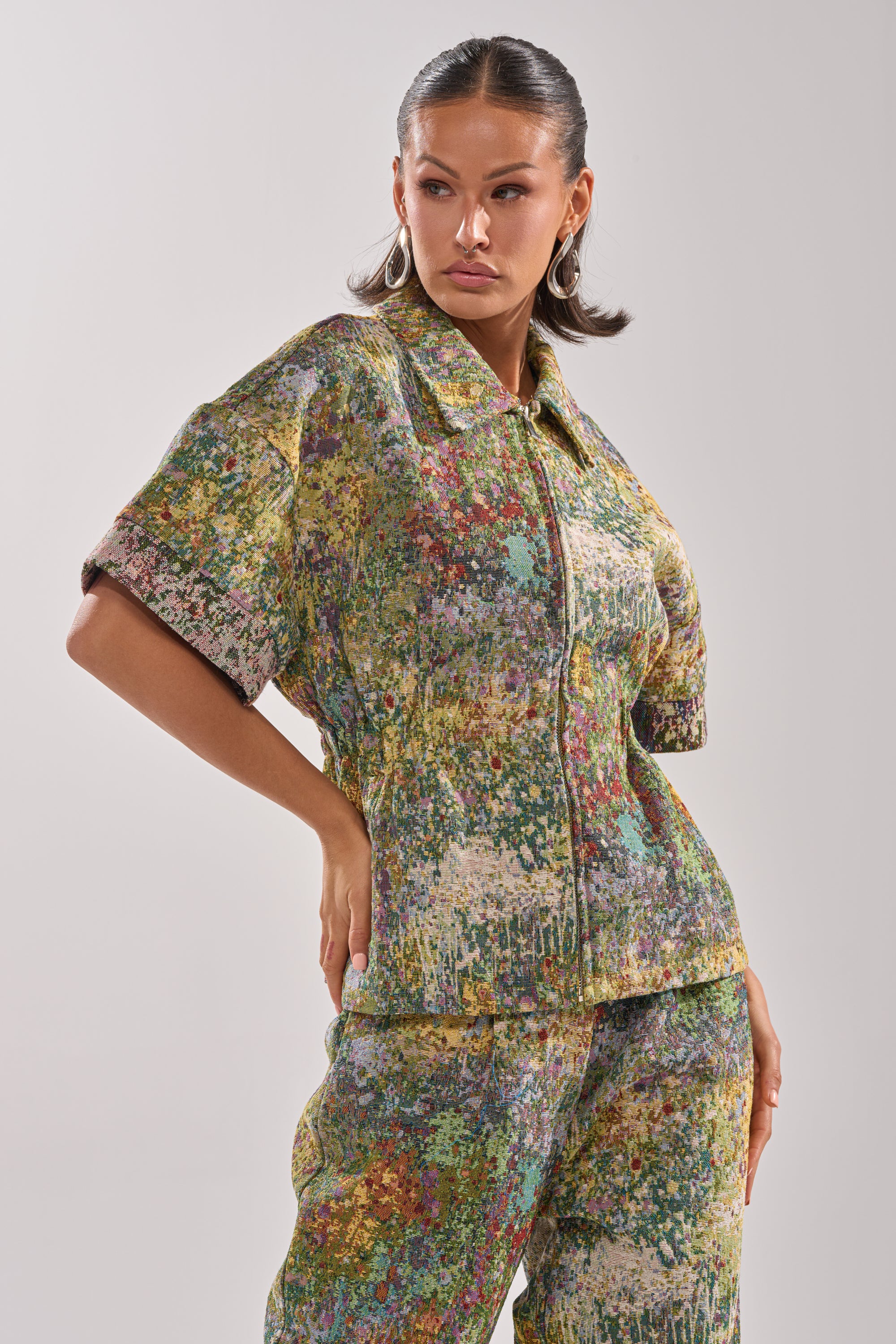 A woman poses confidently in the DEEP IN THE SEA BROCADE BLOUSE, a colorful abstract print top with short sleeves and a zip-up front, paired with matching pants against a plain light background.