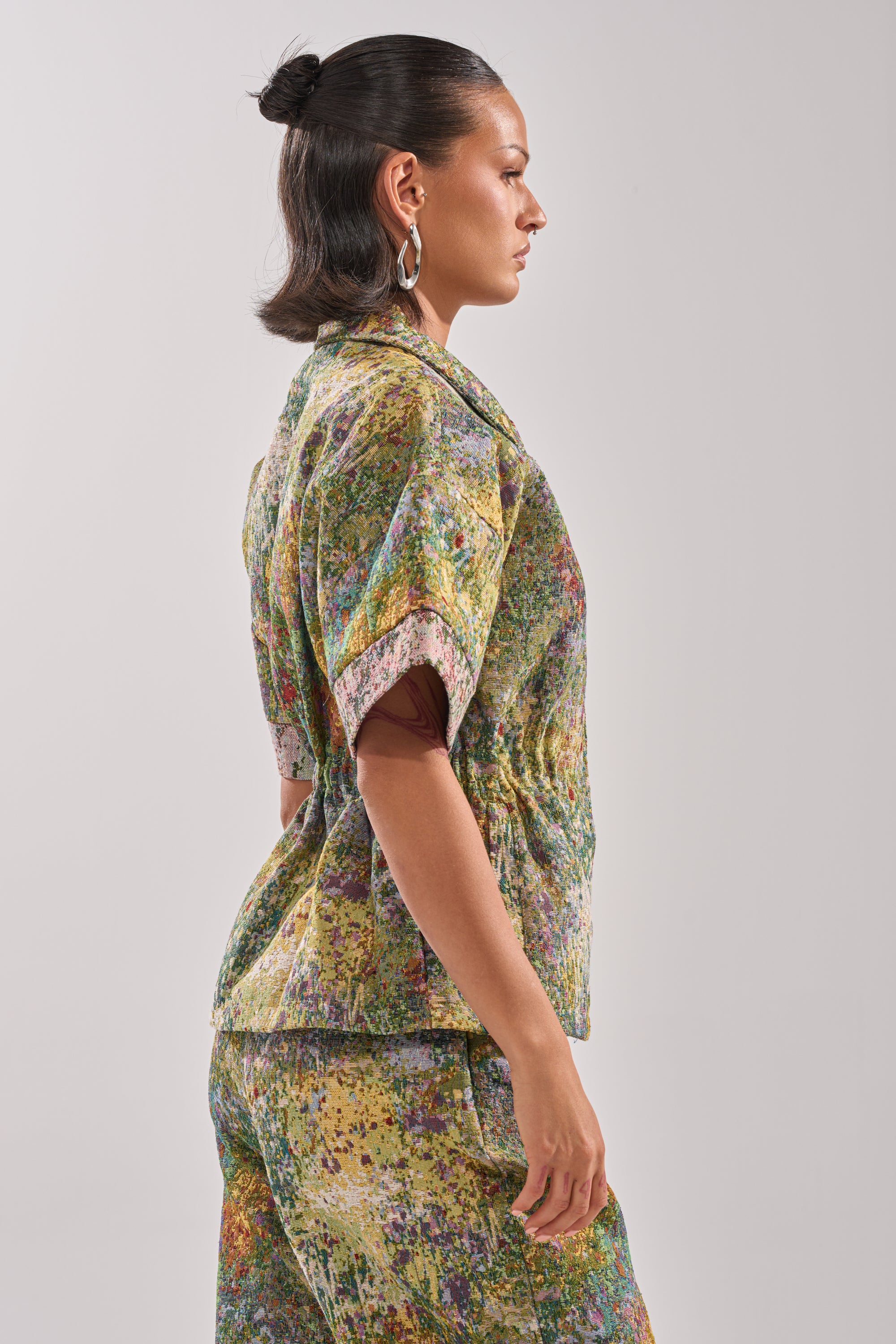 A woman with dark hair in a bun wears the DEEP IN THE SEA BROCADE BLOUSE and matching pants, standing in profile against a plain, light background.