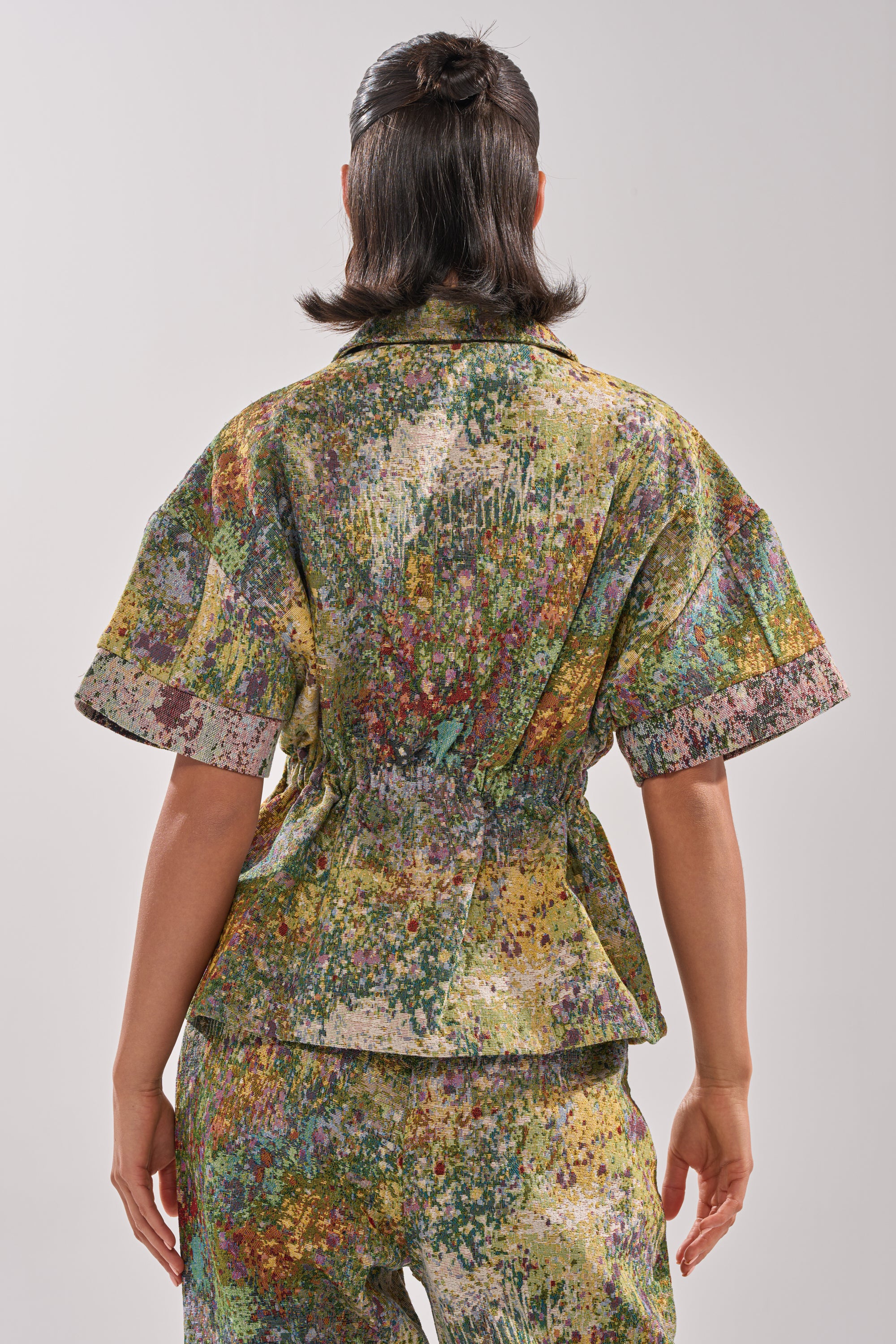 A person faces away, wearing the DEEP IN THE SEA BROCADE BLOUSE and matching pants in a multicolored abstract paint-splatter print. The outfit has a relaxed fit, and their dark hair is styled in a half-updo.