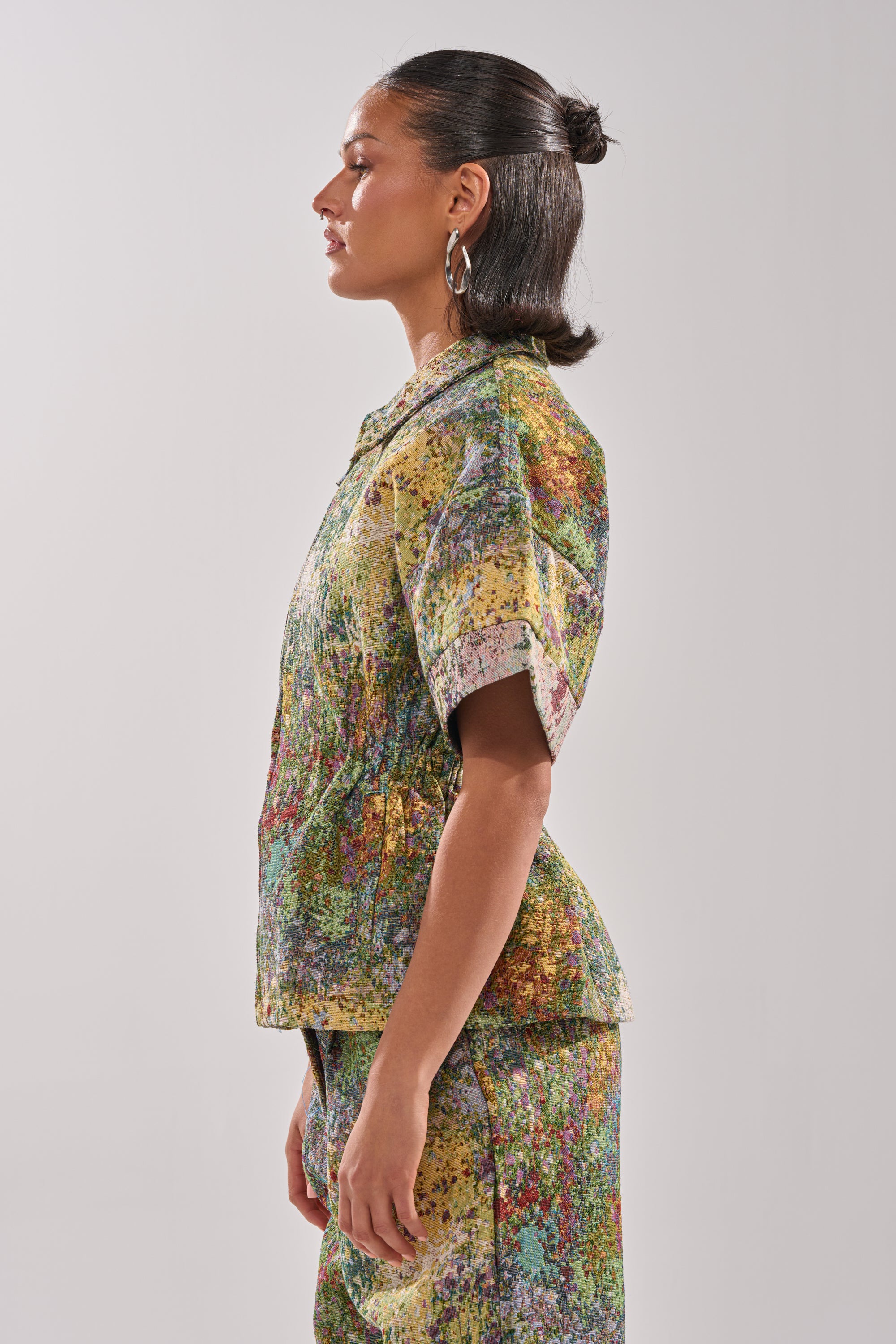 A woman in profile wears the DEEP IN THE SEA BROCADE BLOUSE and matching skirt. Her dark hair is styled in a sleek, low updo, complemented by large hoop earrings against a plain light background.