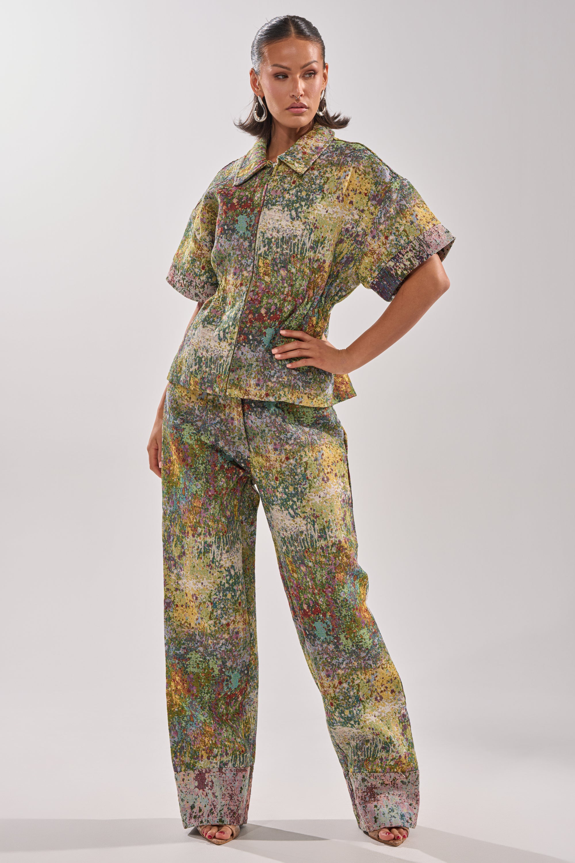 A woman poses confidently in the DEEP IN THE SEA BROCADE BLOUSE with matching wide-leg pants, accessorized with hoop earrings and open-toe heels, set against a plain background.