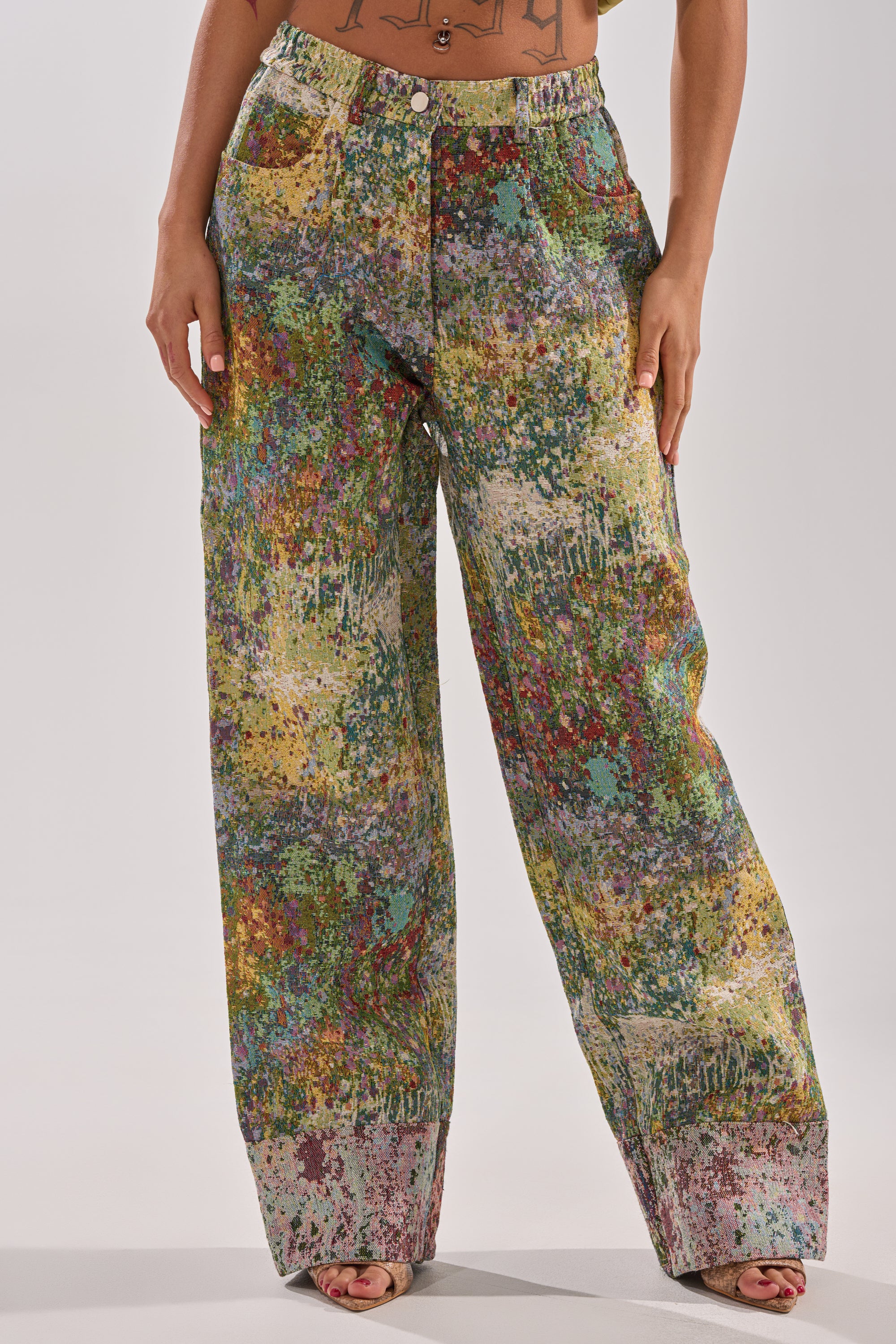 A person wears the DEEP IN THE SEA BROCADE TROUSER, featuring a high-rise, wide-leg silhouette and a colorful abstract paint splatter print, standing with hands at their sides against a plain background.
