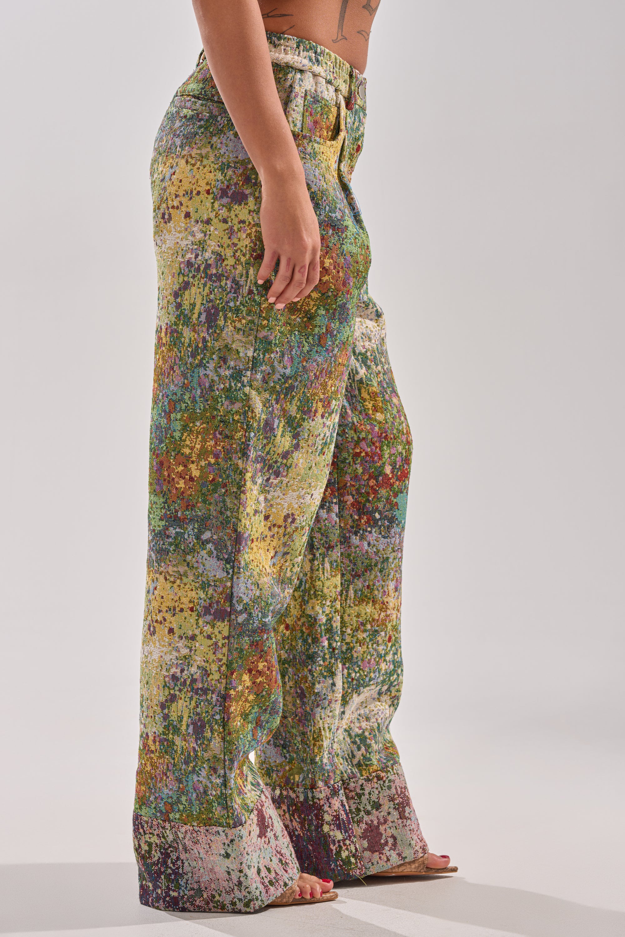 Wearing the DEEP IN THE SEA BROCADE TROUSER—high-rise, wide-leg pants with a colorful abstract floral pattern—a person stands sideways against a plain light background, showing only their midsection to feet and one arm.