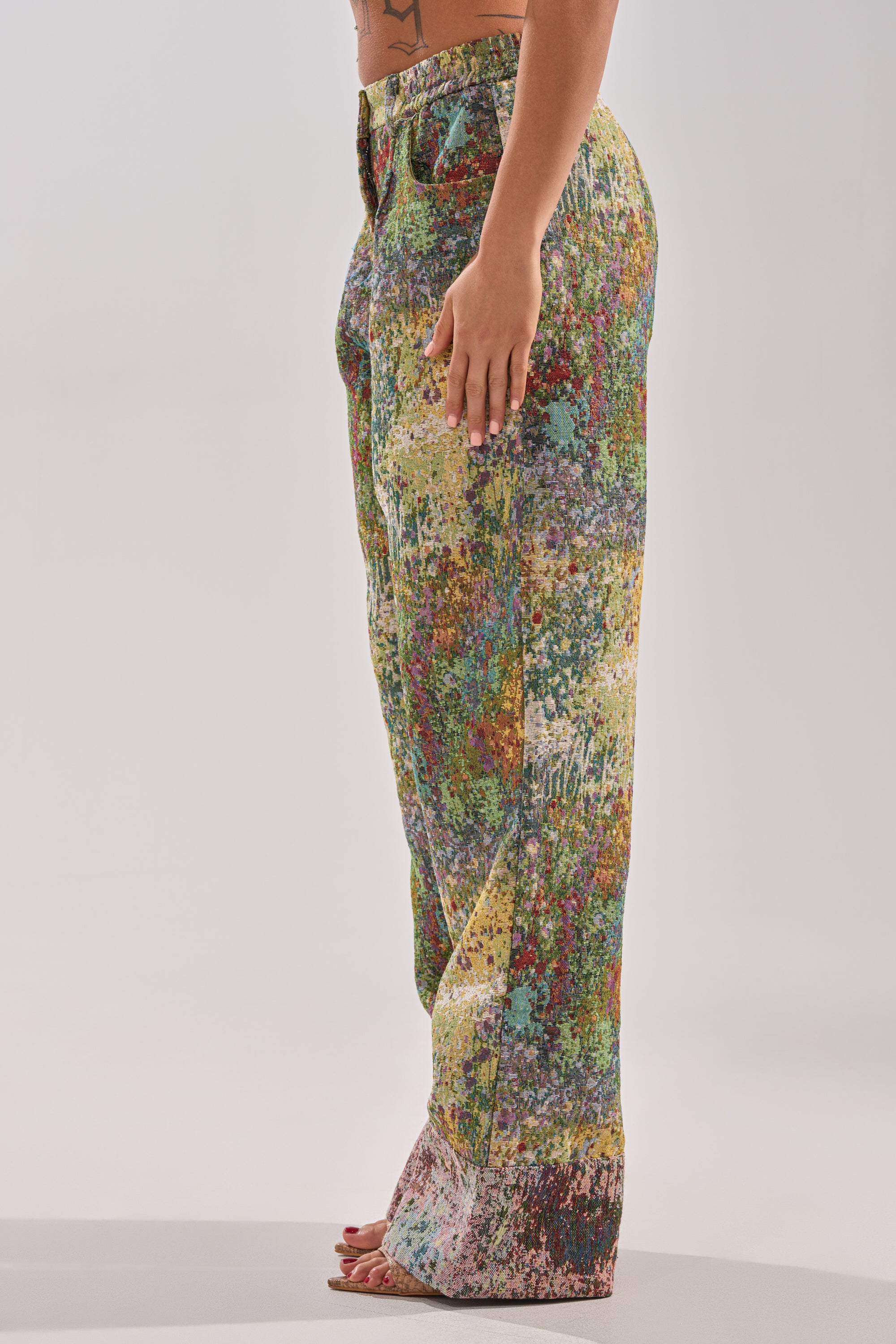 A person stands barefoot on a white background, shown from the side, wearing the DEEP IN THE SEA BROCADE TROUSER—a wide-leg, high-rise pant with a colorful abstract speckled pattern.