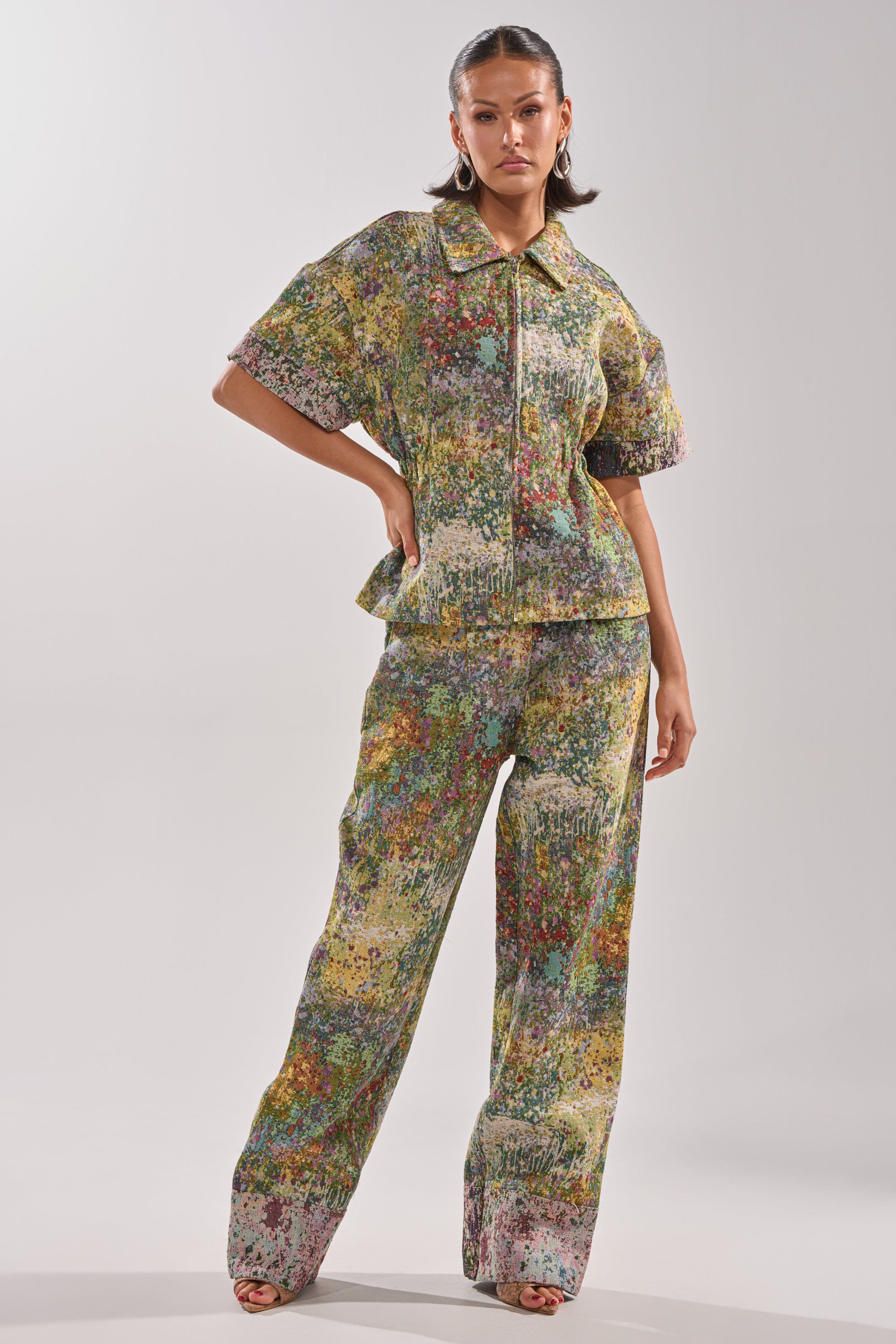 A woman models the DEEP IN THE SEA BROCADE TROUSER, a wide-leg, loose-fitting pant with an abstract multicolored pattern, paired with a collared short-sleeve top. She stands against a plain light backdrop.
