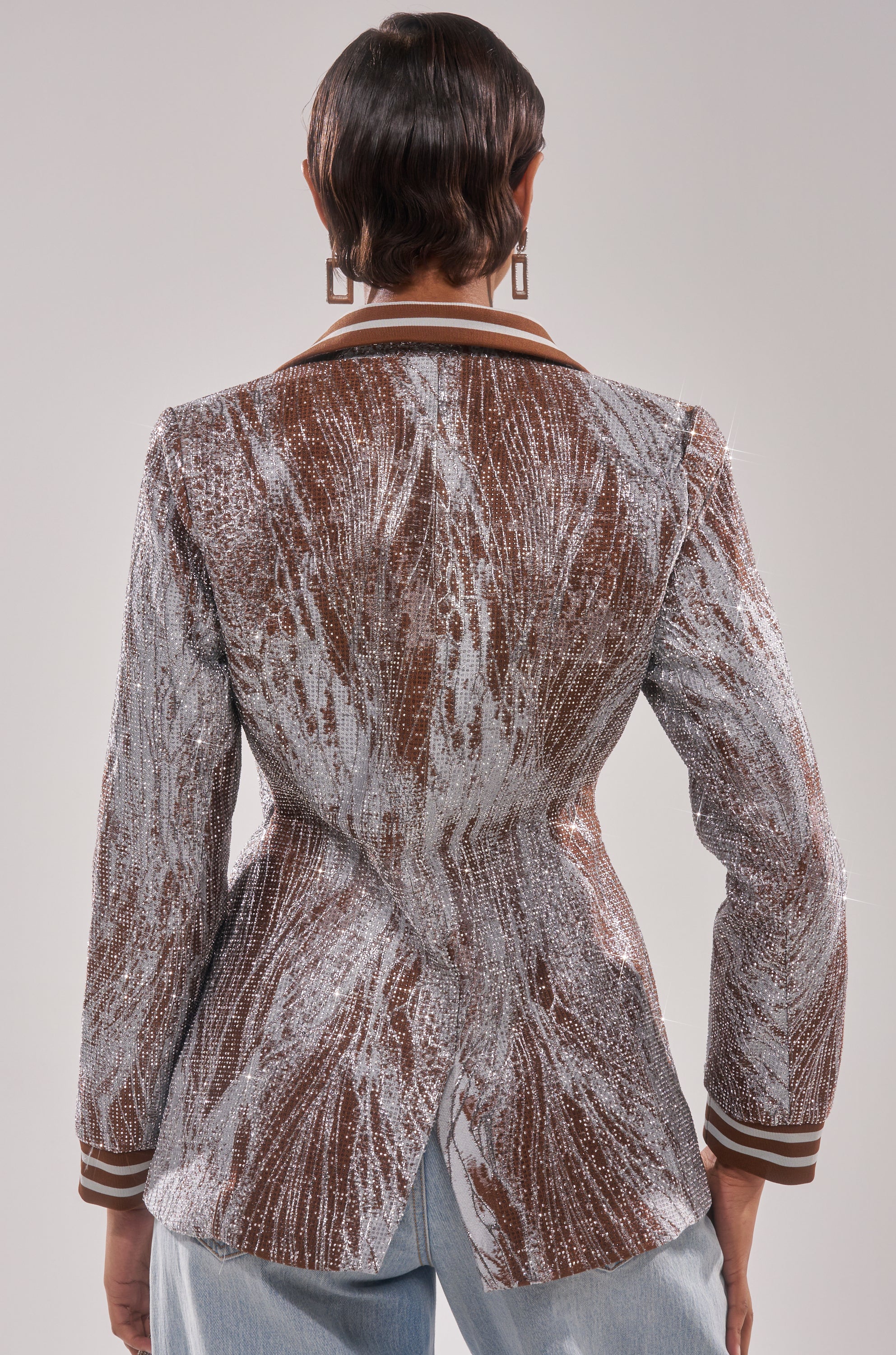 A person with short hair is shown from the back wearing the DASH EMBELLISHED BROCADE BLAZER in brown and silver, styled with light blue jeans. The fitted waist and split hem add flair, completed by geometric earrings for a chic look.
