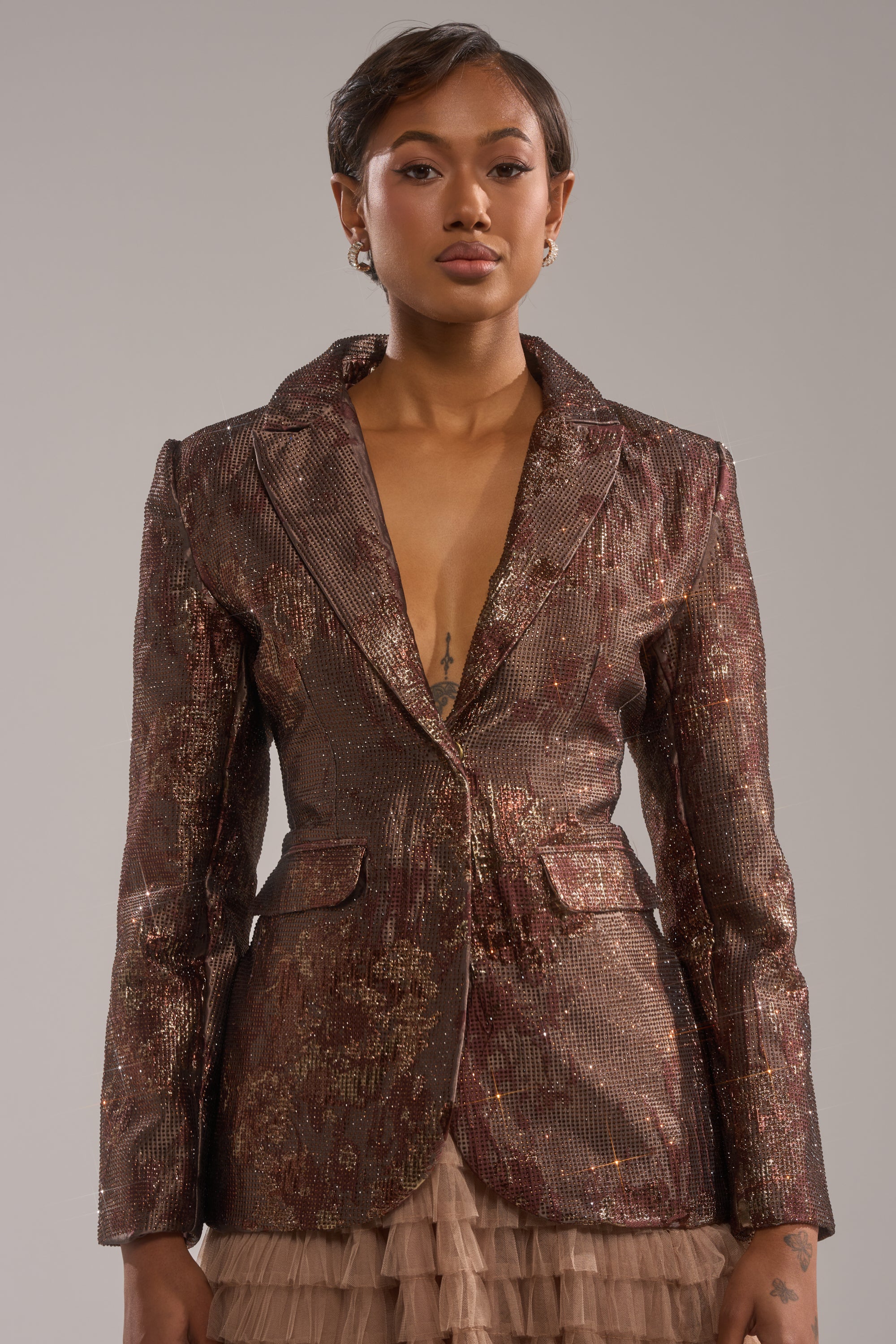 A woman with short hair wears the CELESTE EMBELLISHED BROCADE BLAZER over a ruffled beige skirt, standing against a plain gray background and looking confidently at the camera. 0