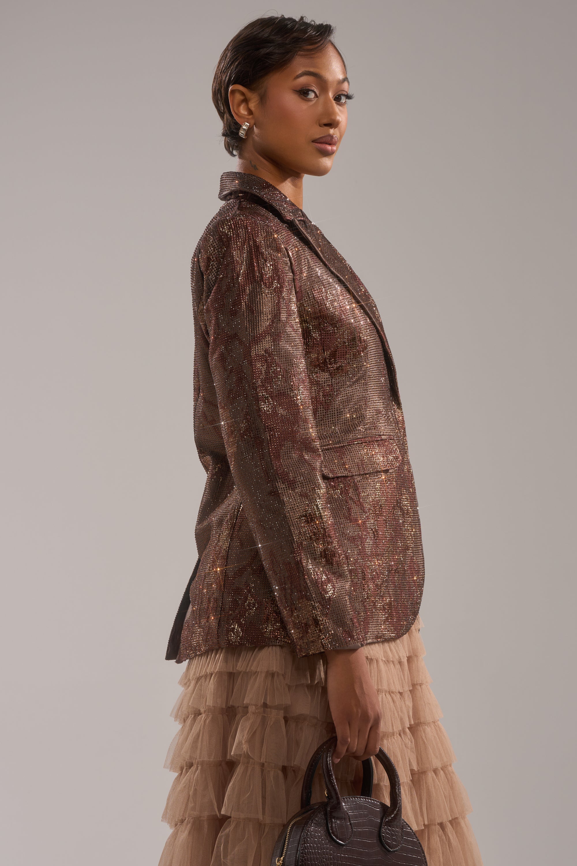 A woman with short hair and statement earrings stands sideways against a plain background, wearing the CELESTE EMBELLISHED BROCADE BLAZER over a beige tiered tulle skirt and holding a dark brown handbag. 2