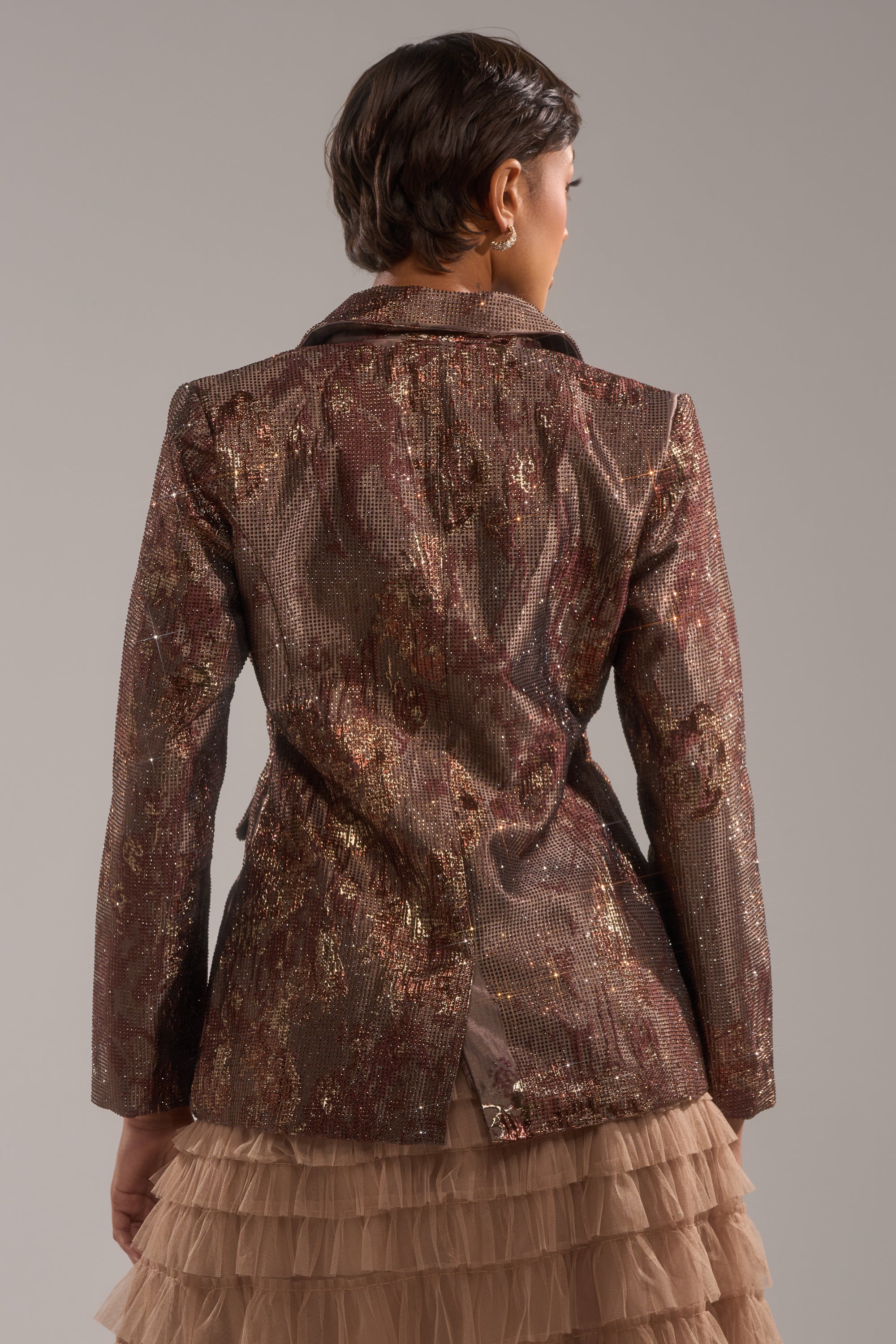 A person with short brown hair is seen from behind in the CELESTE EMBELLISHED BROCADE BLAZER and a layered beige tulle skirt, set against a plain gray background. 3