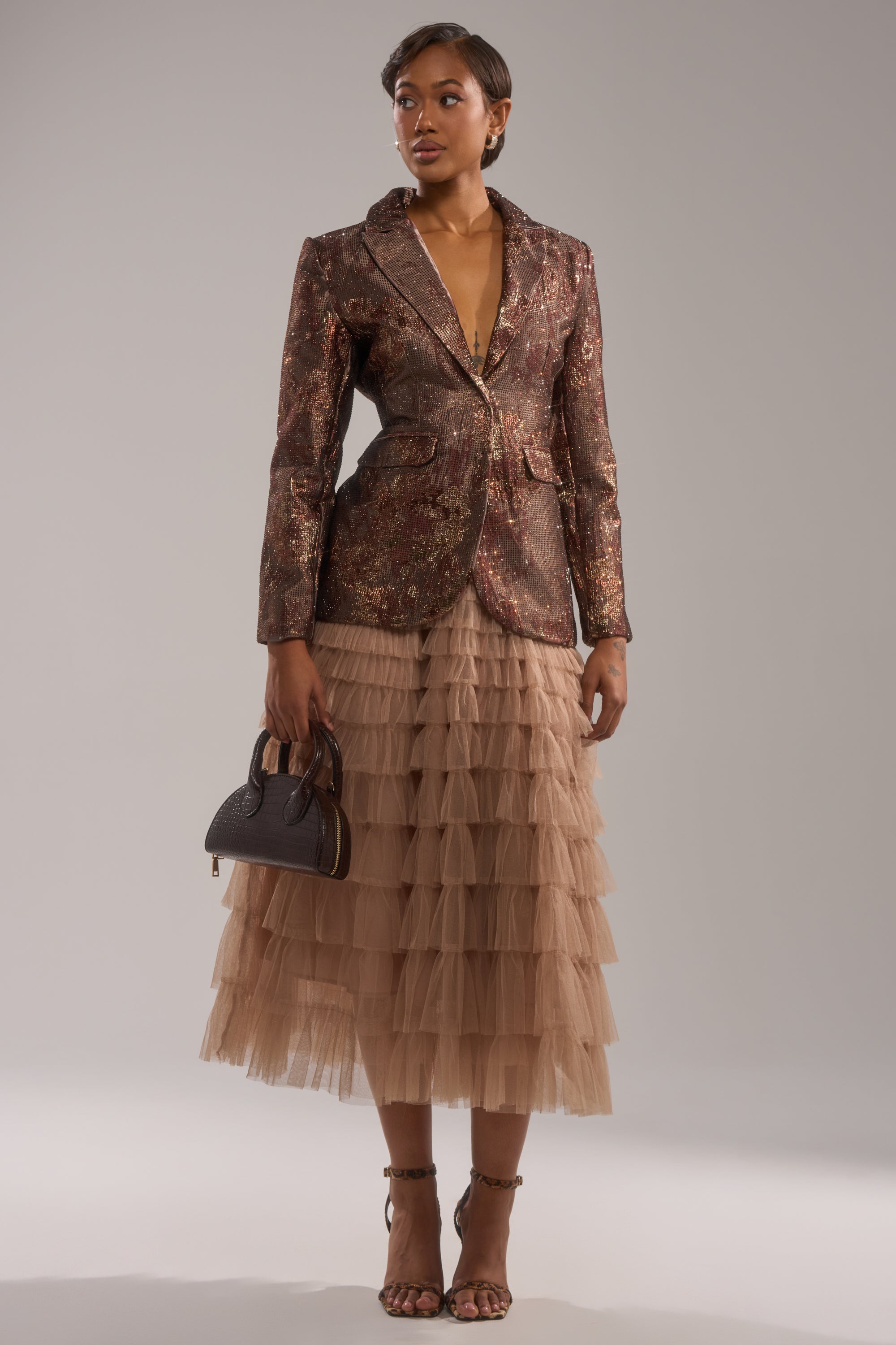 A woman with short hair poses against a plain light background, wearing the CELESTE EMBELLISHED BROCADE BLAZER over a layered beige tulle skirt, styled with strappy heels and a small black handbag. 4