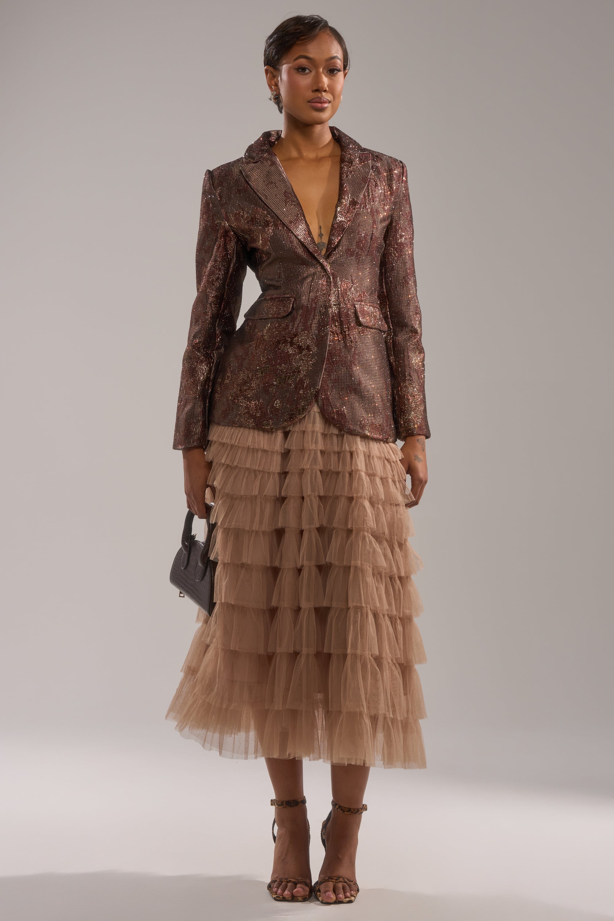 A woman stands confidently in the CELESTE EMBELLISHED BROCADE BLAZER, paired with a layered beige tulle skirt. She holds a small handbag, wears strappy heels, and poses against a plain background with neatly styled hair. 5