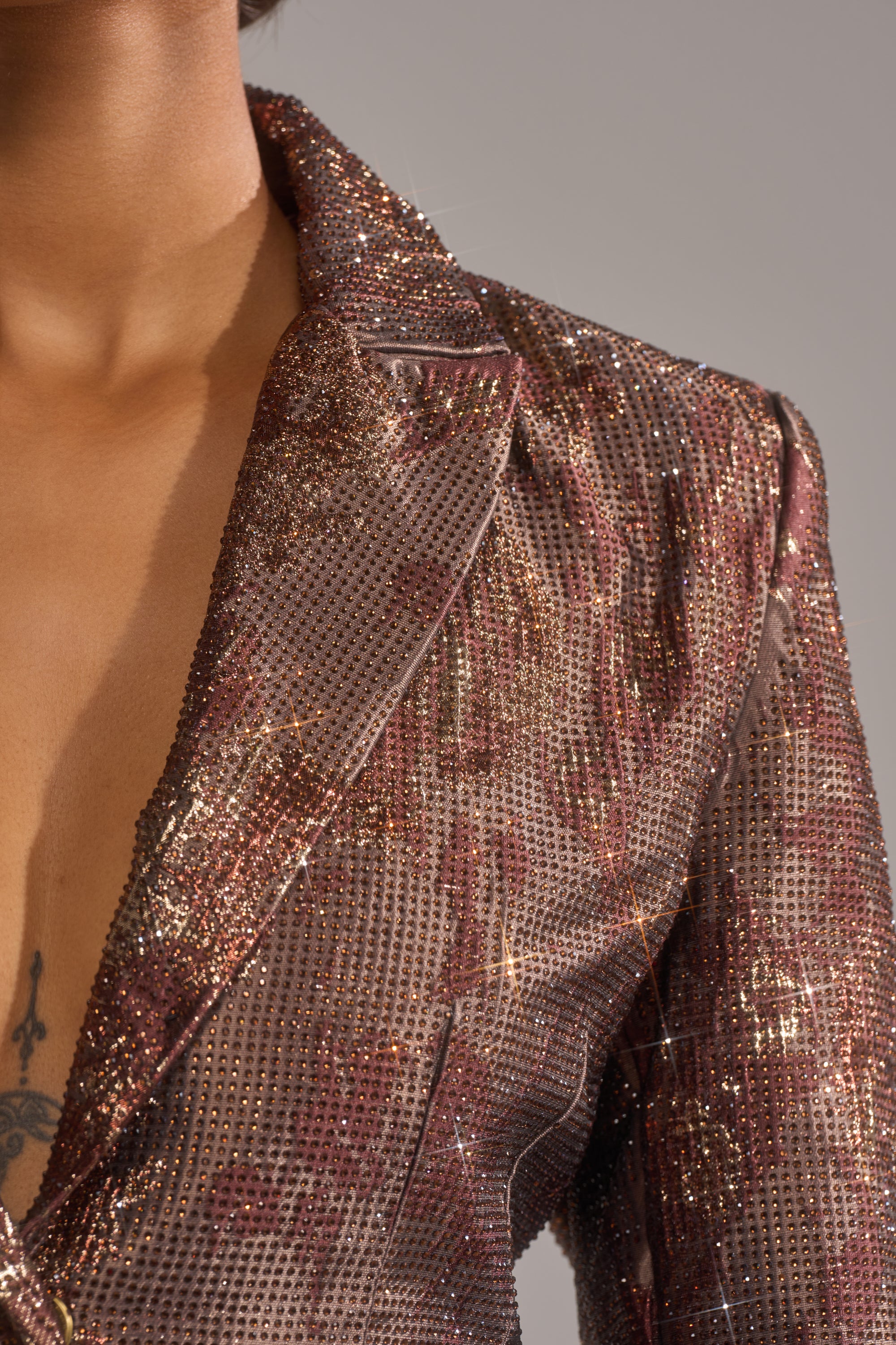 A person wears the CELESTE EMBELLISHED BROCADE BLAZER, sparkling with rhinestones in brown and gold tones. A deep V-neck partially reveals a chest tattoo against a neutral gray background. 6