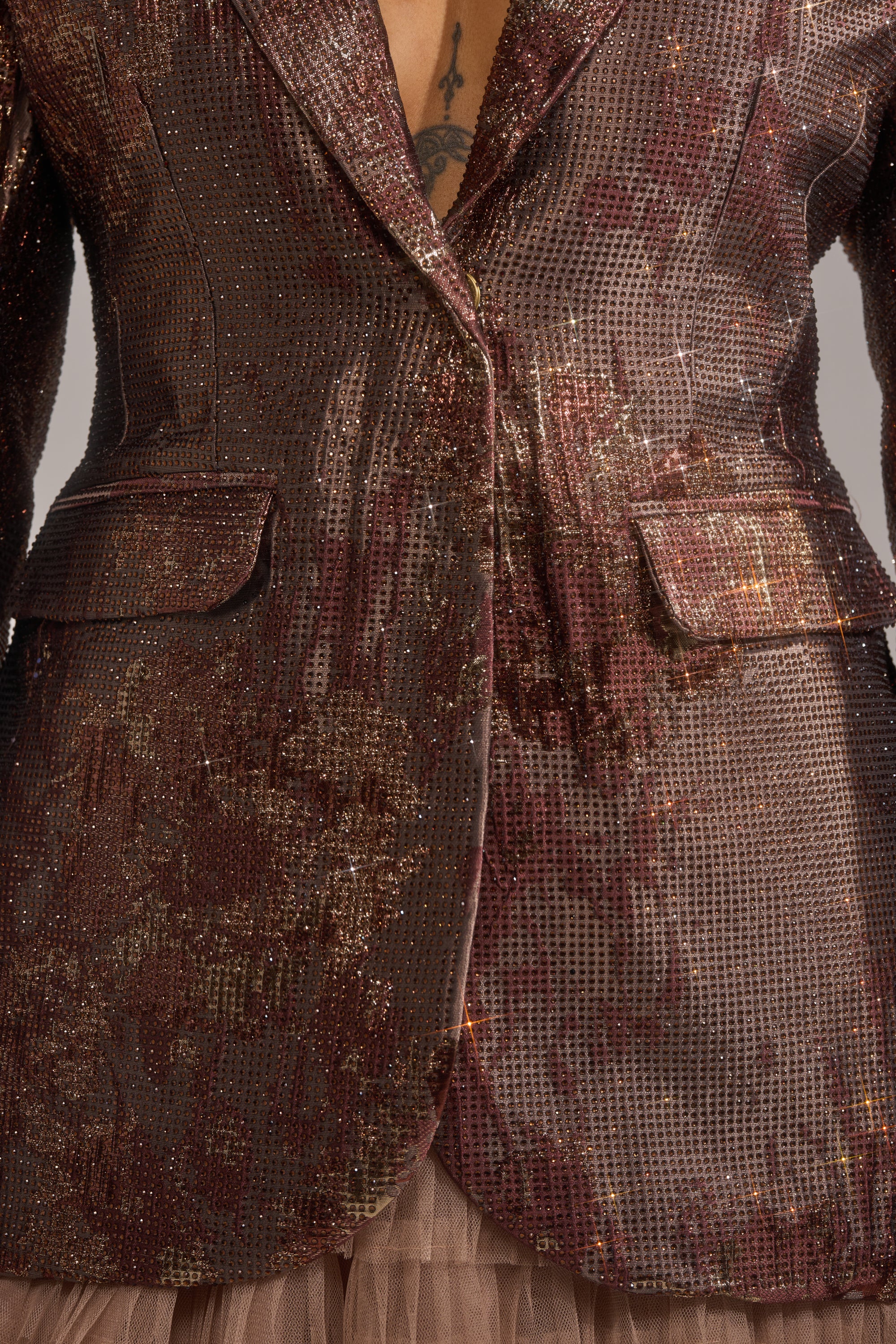 Close-up of a person wearing the CELESTE EMBELLISHED BROCADE BLAZER in shiny dark brown and copper sequins with flap pockets over tulle. The textured blazer reveals part of a chest tattoo. 7