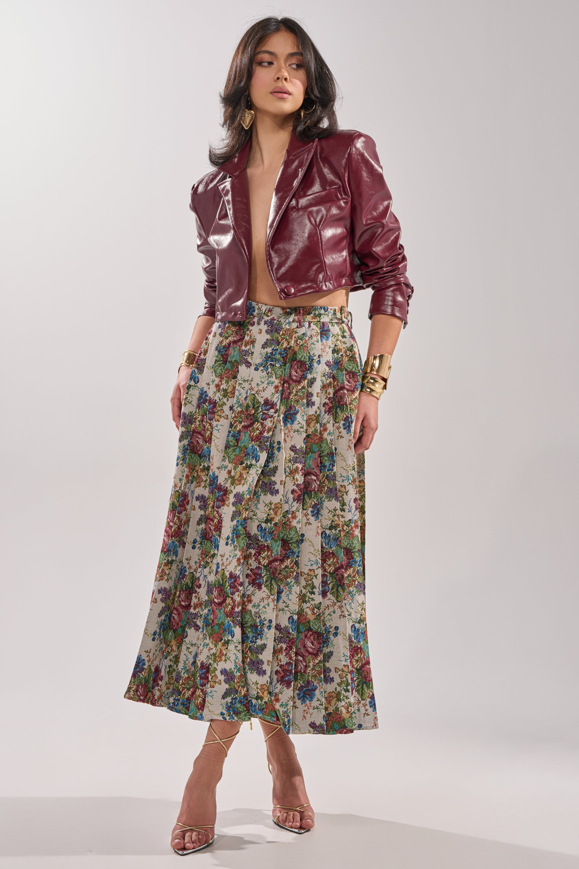 A woman poses in a maroon leather cropped jacket, gold accessories, and the TIFFANY BROCADE MAXI SKIRT with rose print, paired with nude strappy heels against a plain light background. 0