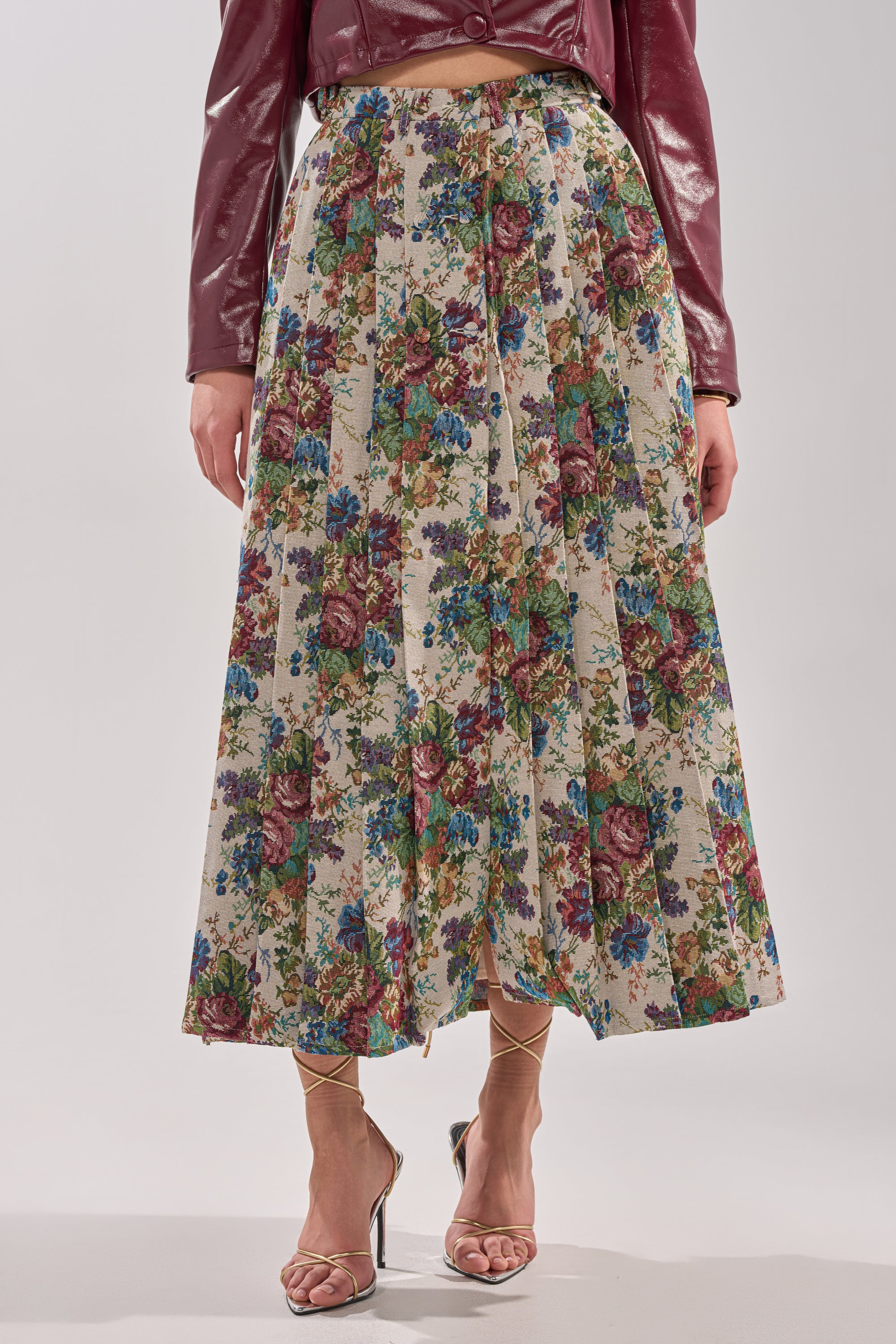 A person wears the TIFFANY BROCADE MAXI SKIRT—a high-waisted, pleated floral cream skirt with red, blue, and green flowers—paired with a maroon shiny jacket and strappy metallic sandals. The upper face is out of frame. 2