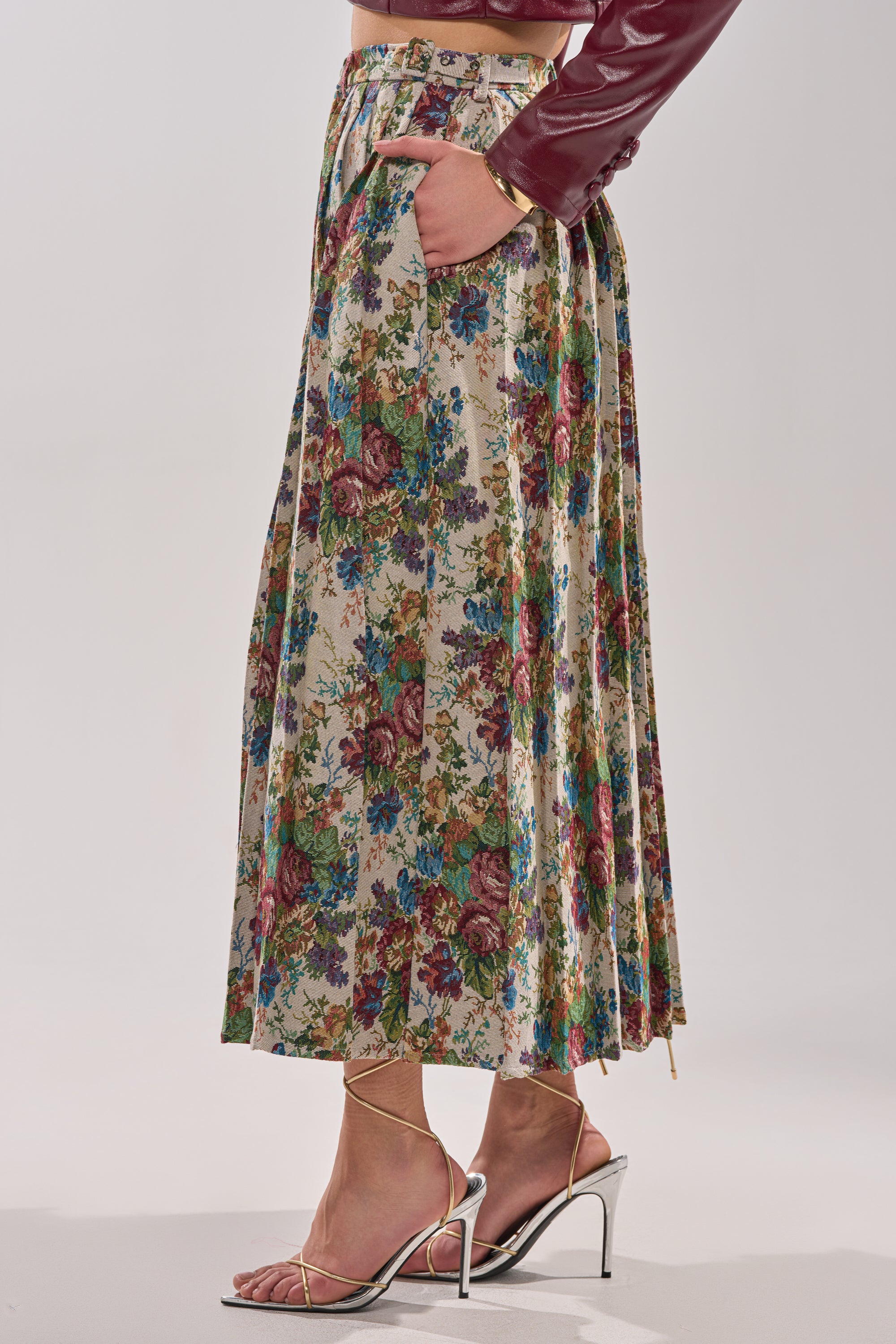 Dressed in the TIFFANY BROCADE MAXI SKIRT with rose print and pockets, a burgundy jacket, and strappy metallic high-heeled sandals, this person stands against a plain light background. 3
