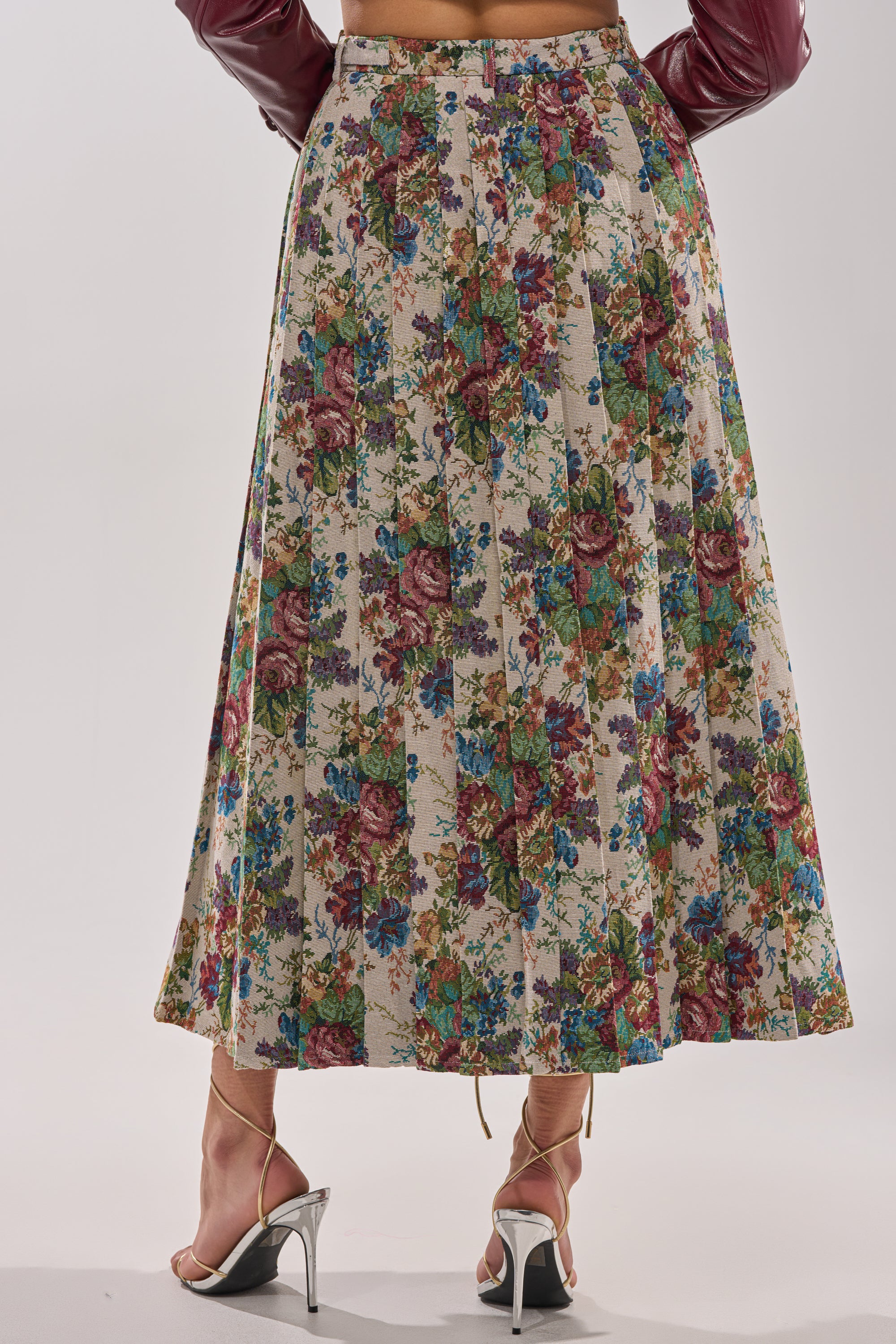 A person is seen from the back wearing the TIFFANY BROCADE MAXI SKIRT, which features a flowy high waist design and a colorful floral pattern, paired with metallic high-heeled sandals. 4
