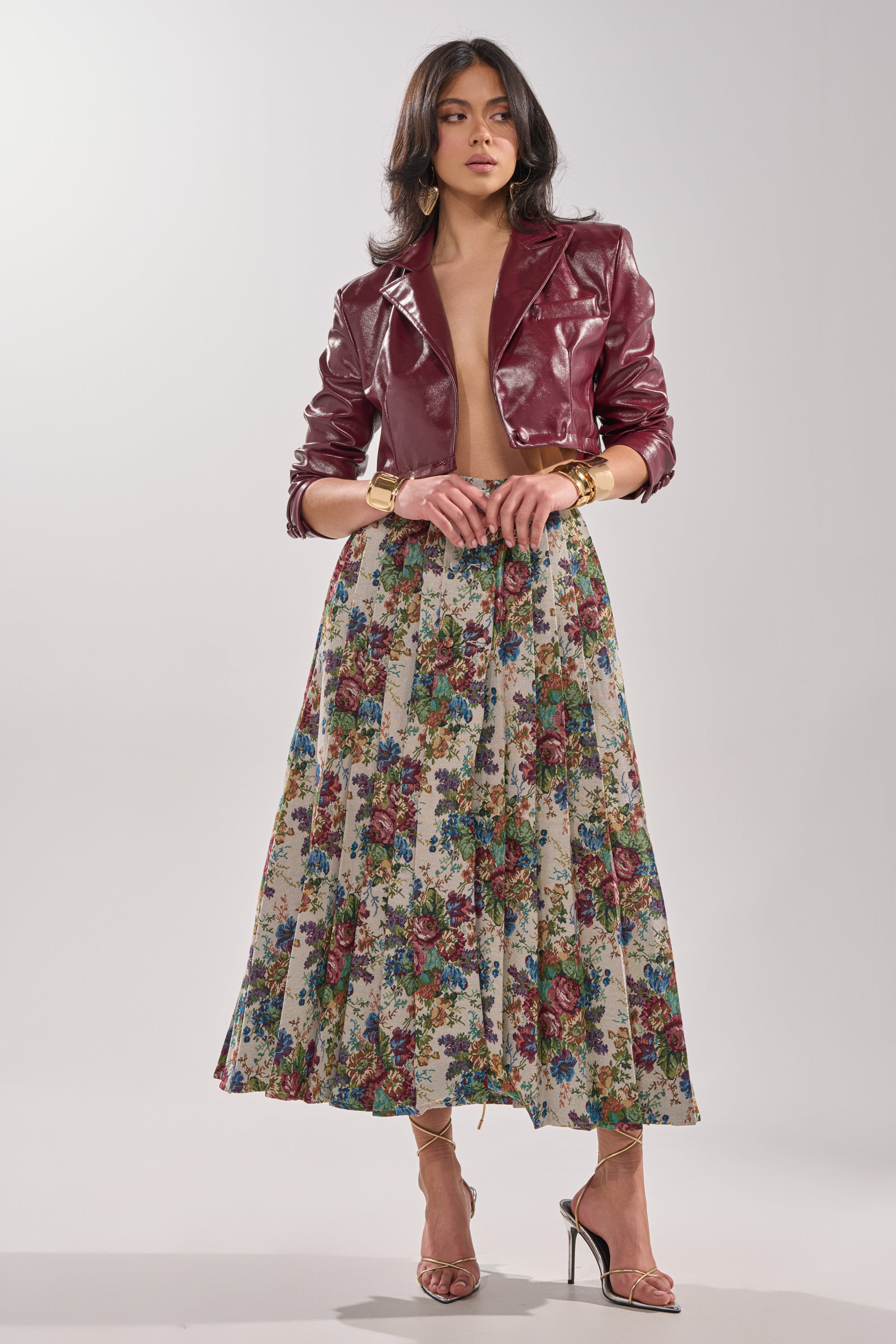 A woman poses in a TIFFANY BROCADE MAXI SKIRT paired with gold jewelry, strappy high heels, and a burgundy faux leather jacket against a plain light background. 5