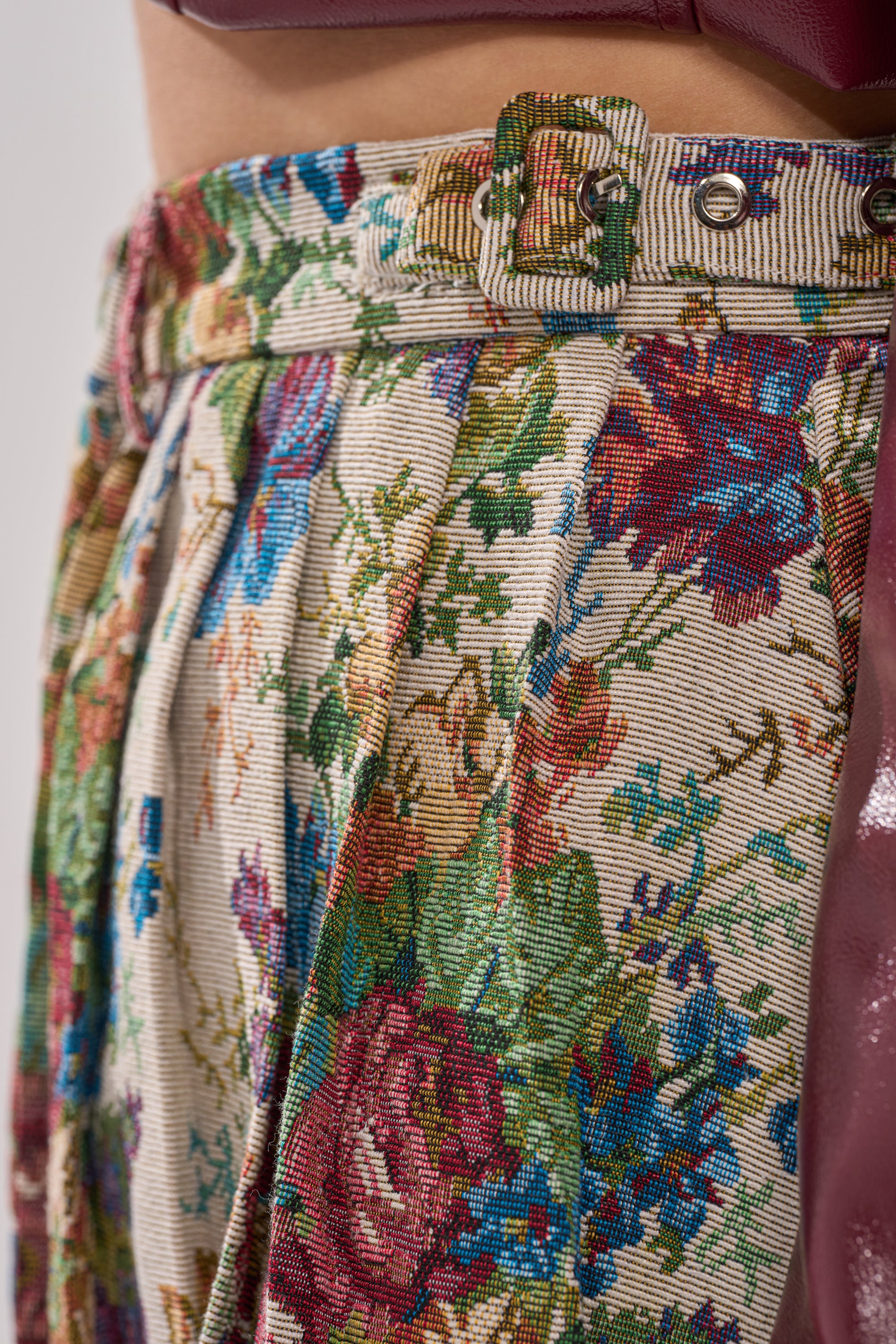 Close-up of the TIFFANY BROCADE MAXI SKIRT featuring a detailed floral tapestry pattern, pleats, and a matching fabric-covered belt. A shiny pink garment appears partially on the right side. 6