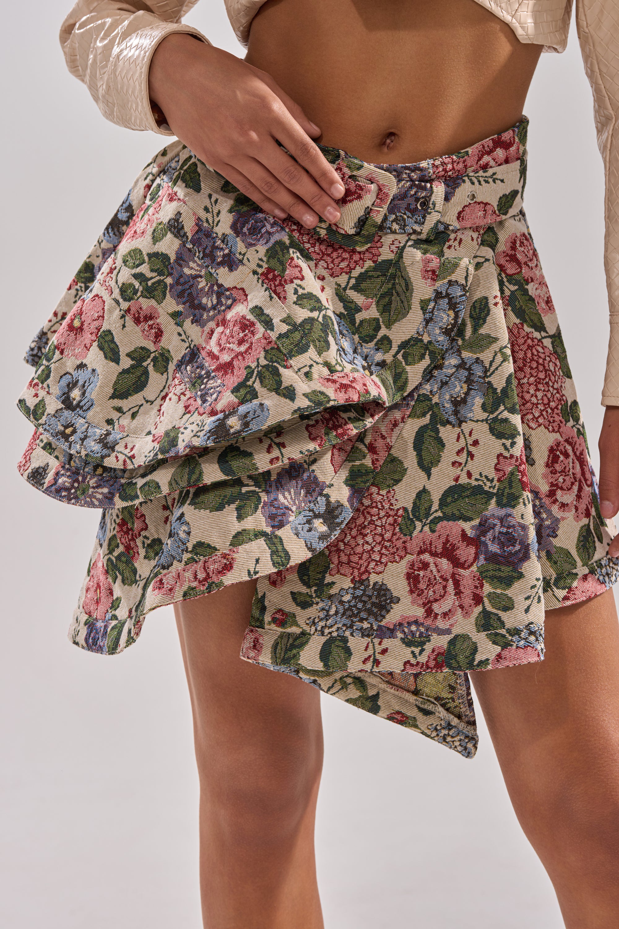 Someone is wearing a cream long-sleeve top paired with the GIVE ME FLOWERS MINI SKIRT—a high-waisted, vintage-inspired floral mini skirt featuring an asymmetrical ruffle layer and matching belt—against a plain, light background.