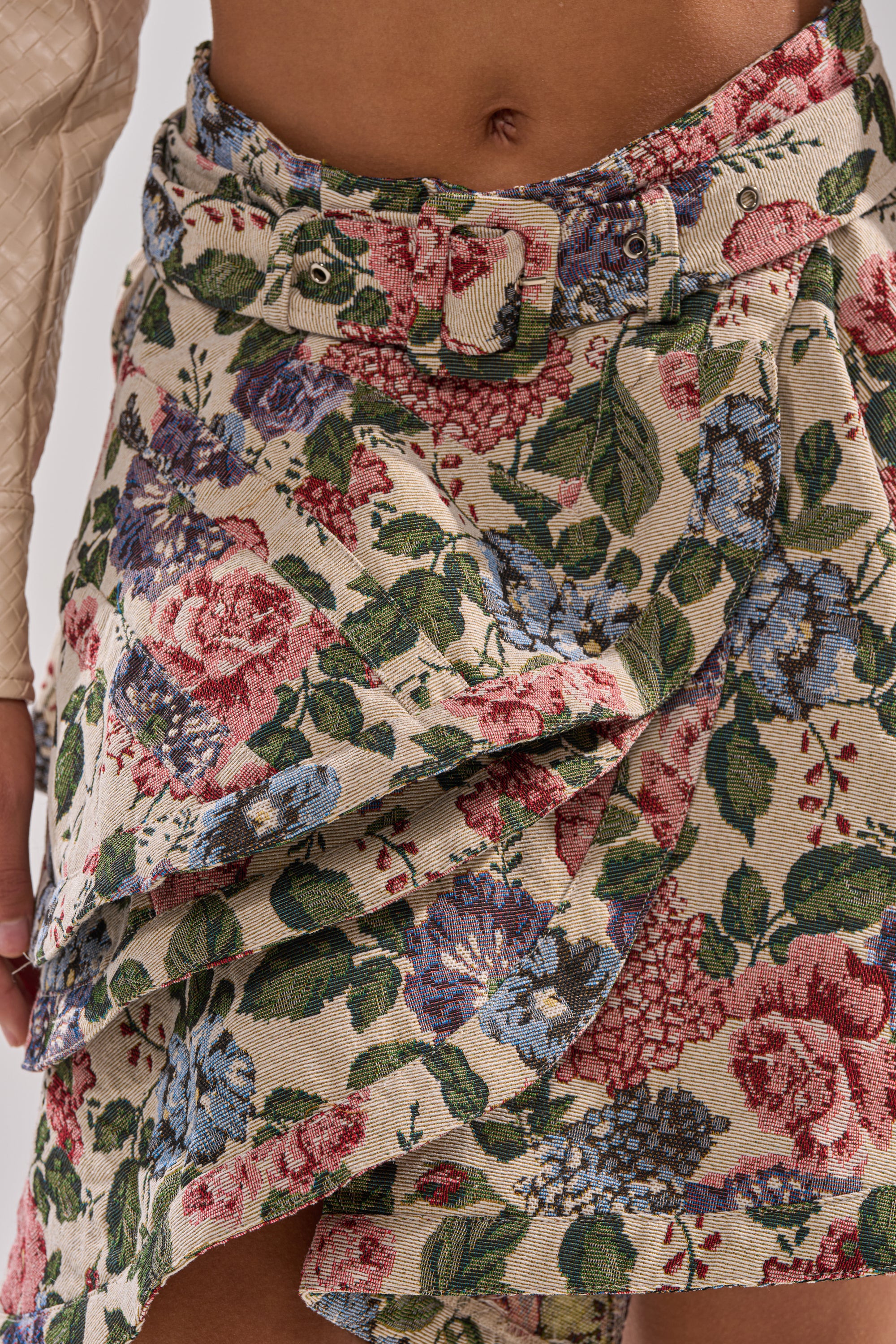 Close-up of a person wearing the GIVE ME FLOWERS MINI SKIRT, a vintage-style, high-rise mini with layered ruffles and matching belt. Features red, blue, and green botanical designs on a beige background.