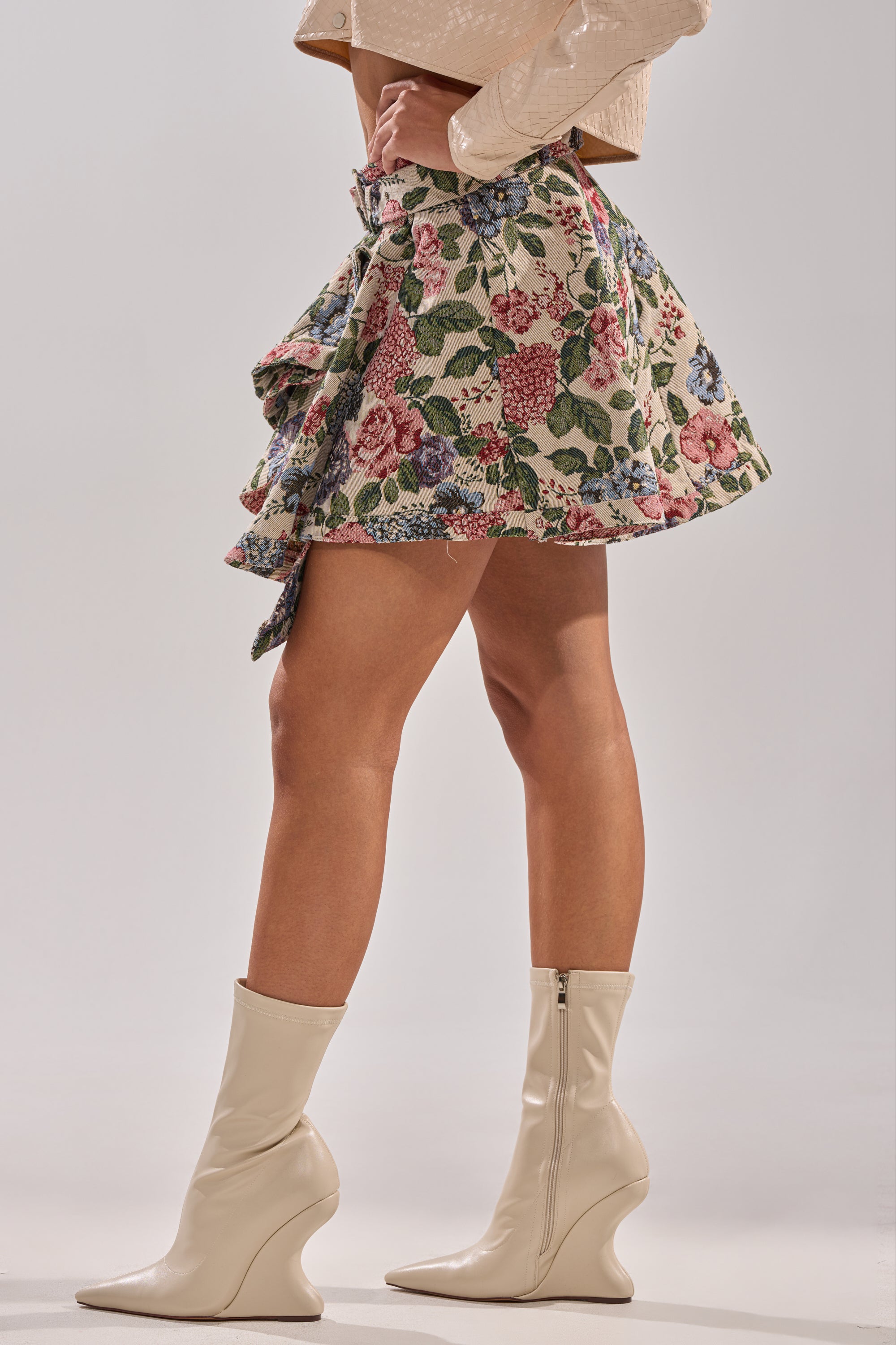 Wearing the GIVE ME FLOWERS MINI SKIRT, a beige textured long-sleeve top, and cream wedge-heeled ankle boots, a person poses with one hand on their hip against a plain background.