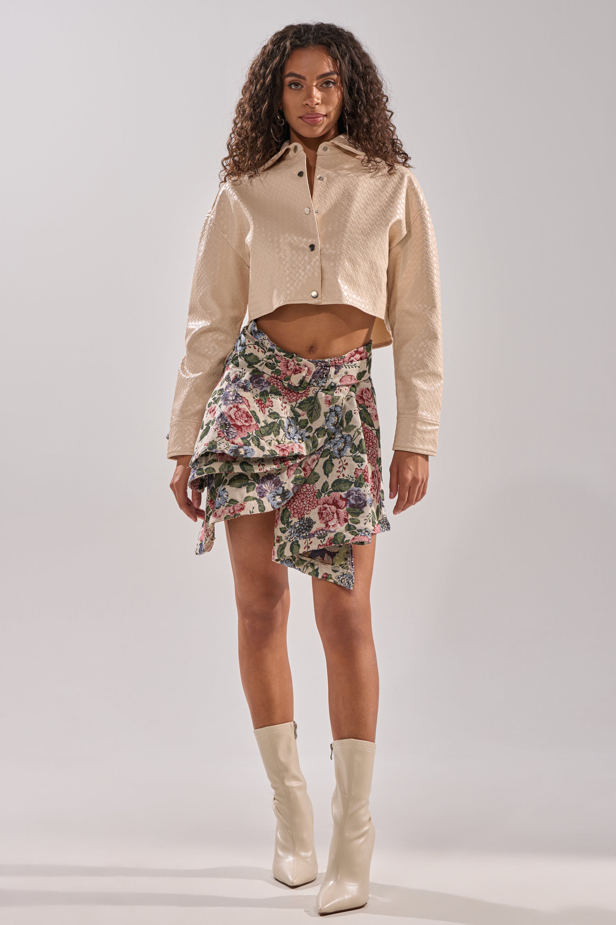 A woman with long curly hair and a neutral expression stands facing forward, wearing the GIVE ME FLOWERS MINI SKIRT, a cropped beige button-up jacket, and cream knee-high boots against a plain light background.