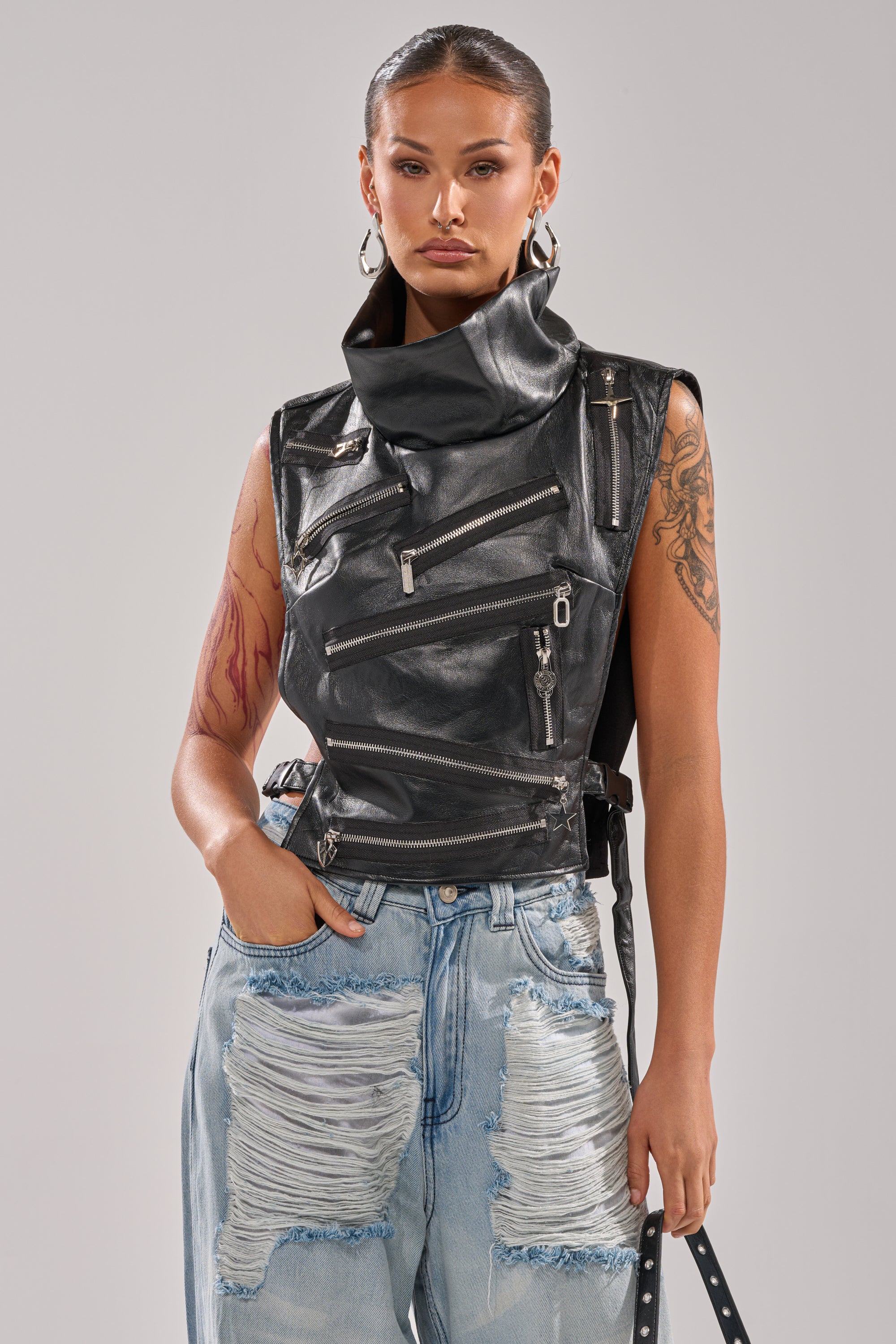 A woman with tattoos and hoop earrings wears the ZIP ME UP TURTLENECK TOP IN BLACK with ripped light blue jeans, posing confidently against a plain gray background.