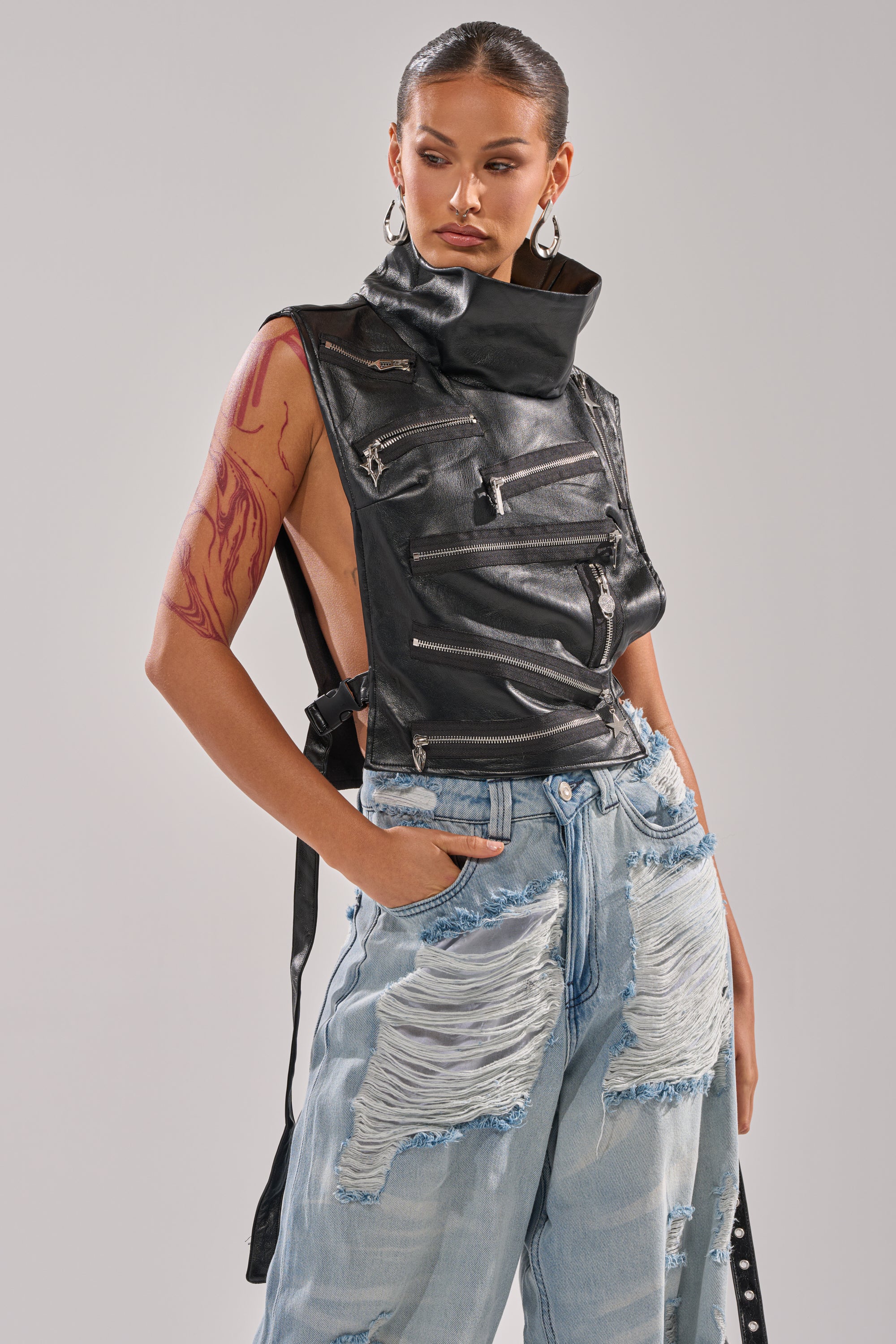 Model wears the ZIP ME UP TURTLENECK TOP IN BLACK—cropped faux leather with multiple zippers, buckled straps, and open sides—styled with distressed jeans, hoop earrings, a red arm tattoo, and slicked-back hair.