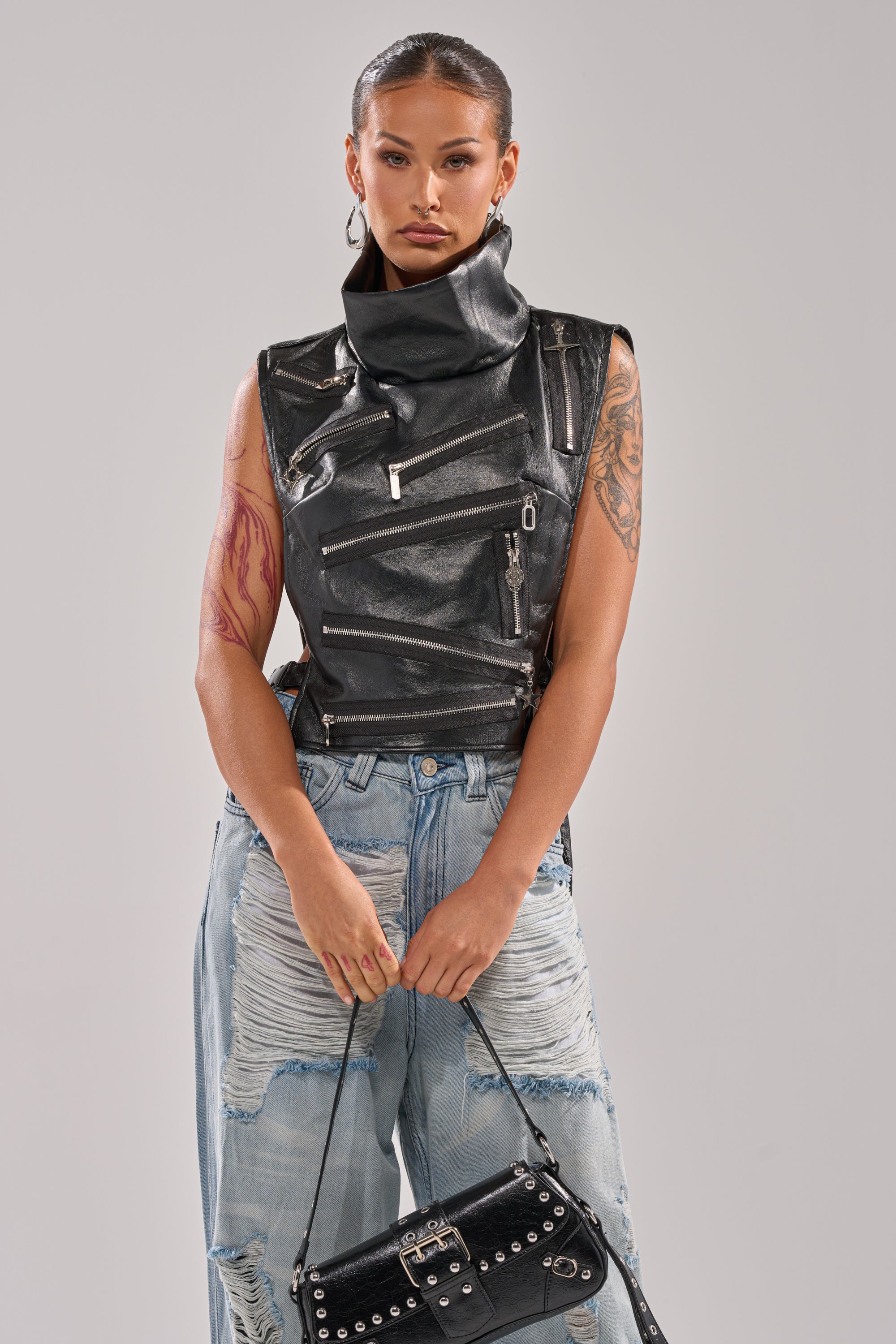 Wearing the ZIP ME UP TURTLENECK TOP IN BLACK with multiple zippers, paired with light blue ripped jeans and holding a black studded handbag, a person stands against a plain light background.