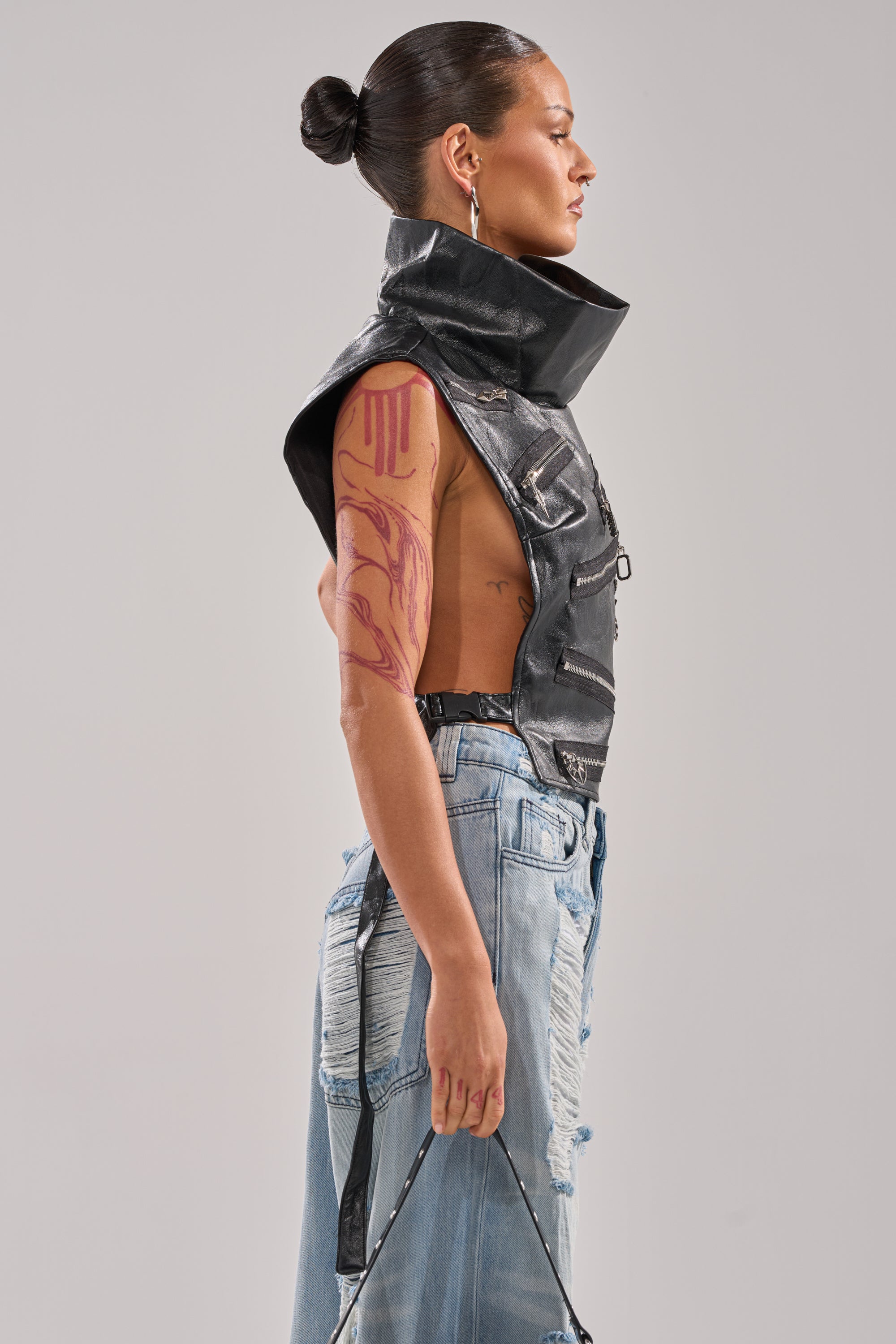 A woman with slicked-back hair stands in profile wearing the ZIP ME UP TURTLENECK TOP IN BLACK and distressed blue jeans. She has a red abstract tattoo on her upper arm and holds an adjustable buckled strap in her hand.