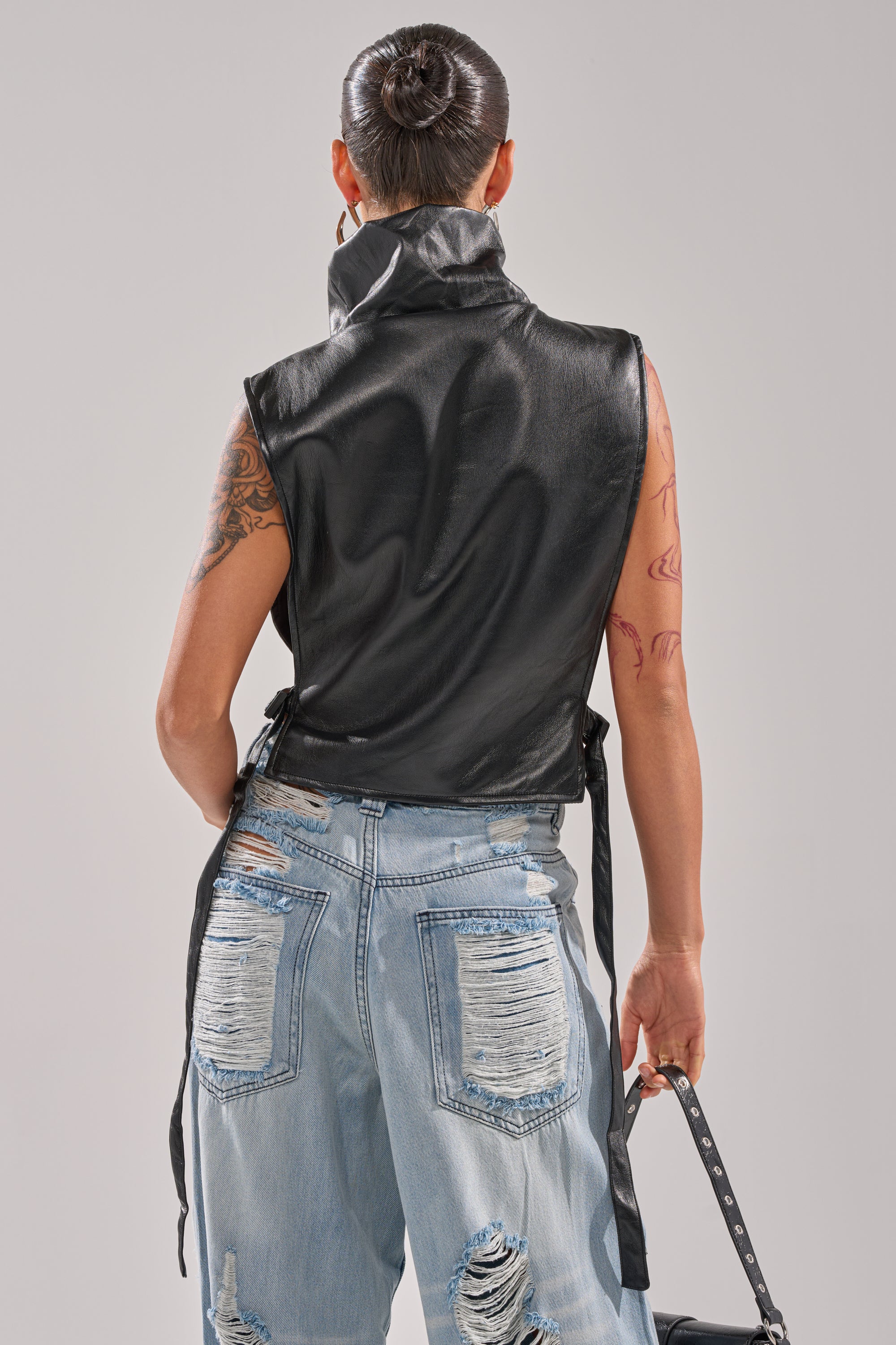 A person with a sleek bun wears the ZIP ME UP TURTLENECK TOP IN BLACK and distressed light blue jeans. With their back to the camera, arm tattoos are visible as they hold a small black bag with adjustable buckled straps.