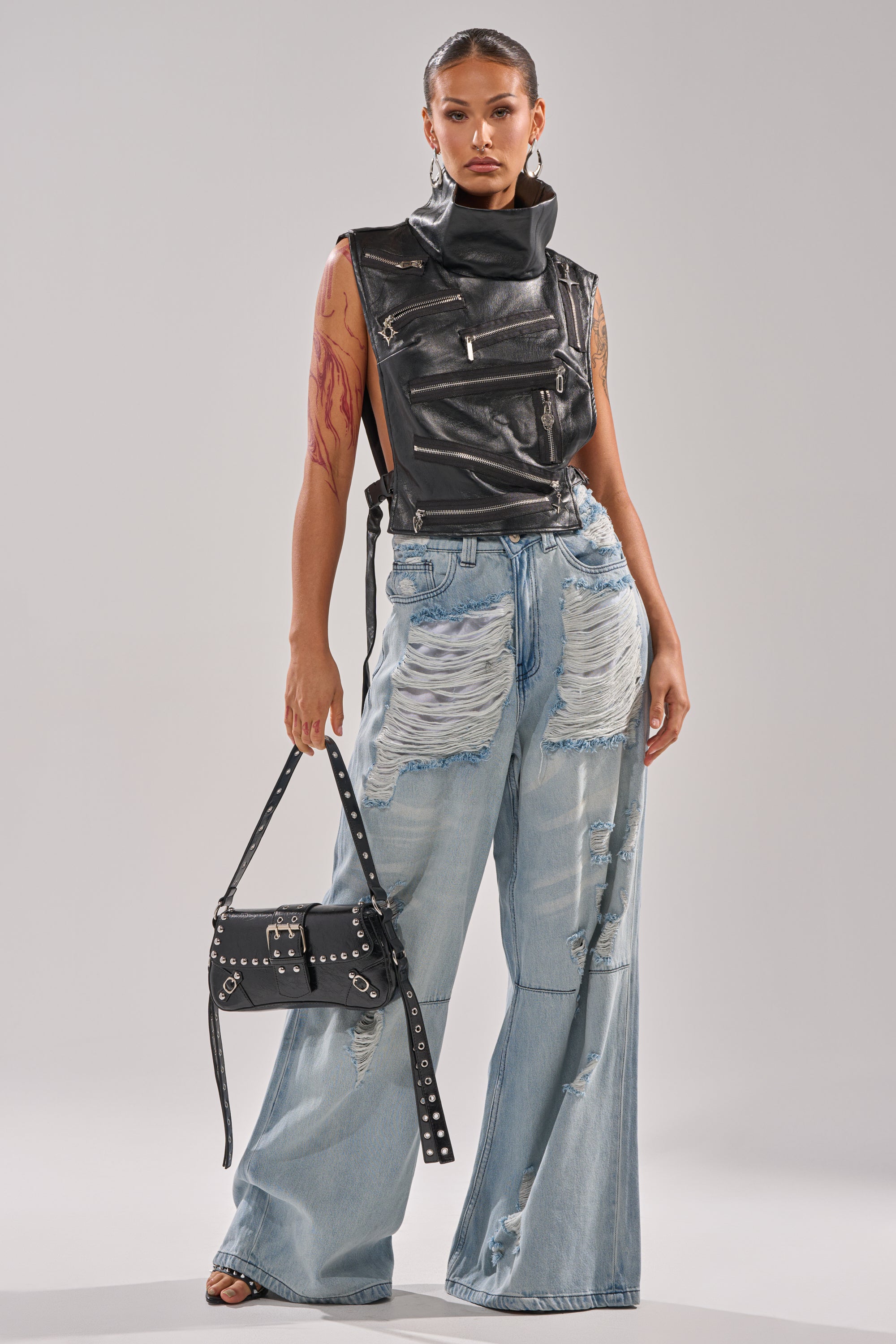 A person poses confidently in the ZIP ME UP TURTLENECK TOP IN BLACK, paired with distressed wide-leg light blue jeans and a black studded handbag for an edgy, fashion-forward look.