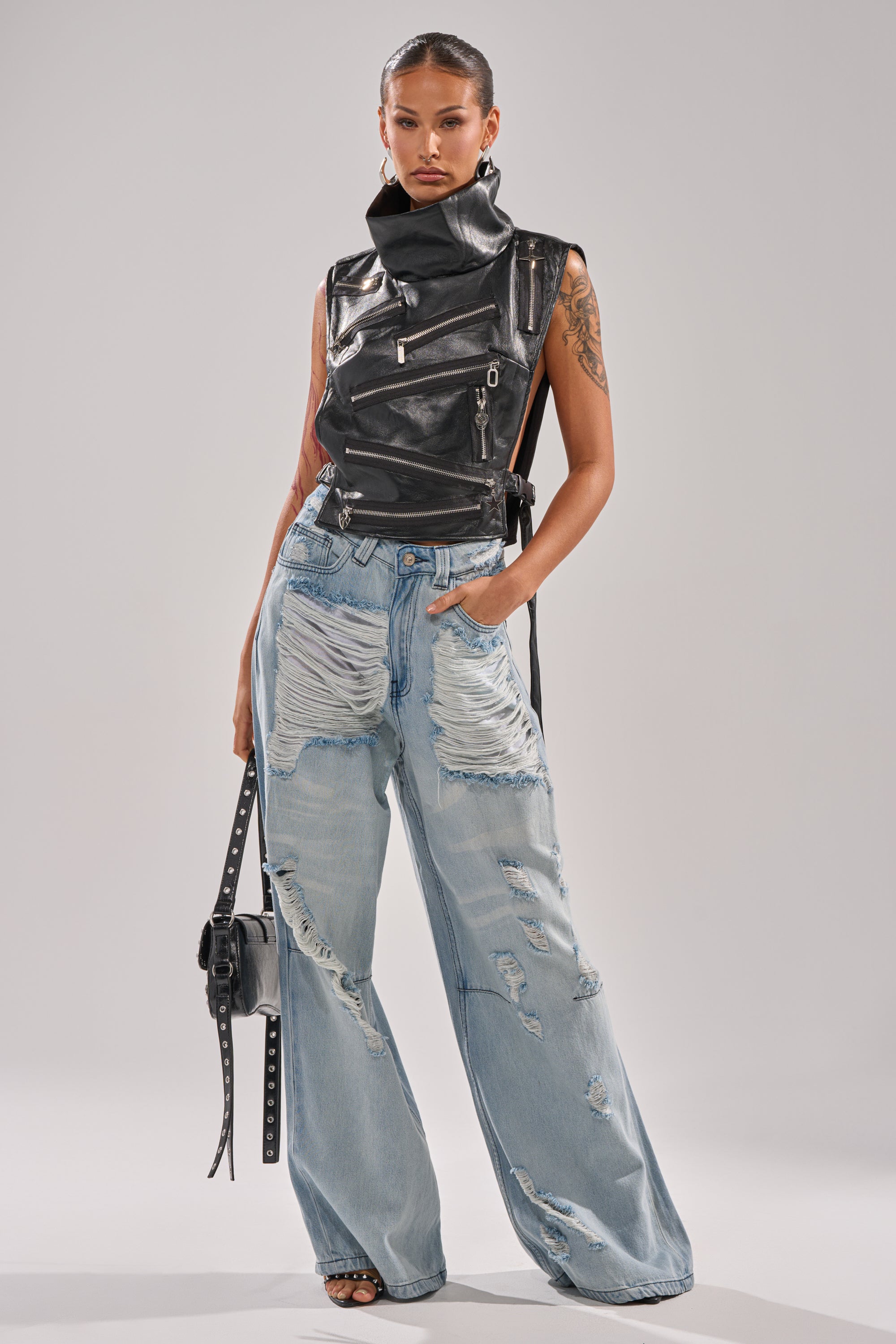 A woman with slicked-back hair and arm tattoos poses confidently in the ZIP ME UP TURTLENECK TOP IN BLACK, paired with distressed light blue wide-leg jeans and a black studded handbag.