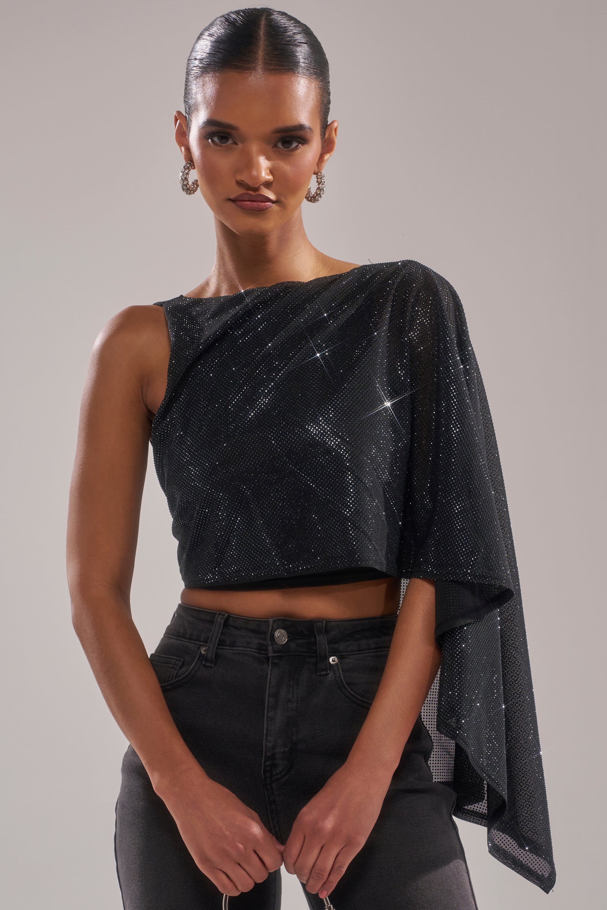 A woman with slicked-back hair and hoop earrings poses against a plain background, wearing the TOO HOT FOR YOU RHINESTONE MESH TOP featuring sparkling black mesh with one draped sleeve, paired with high-waisted black jeans. 0