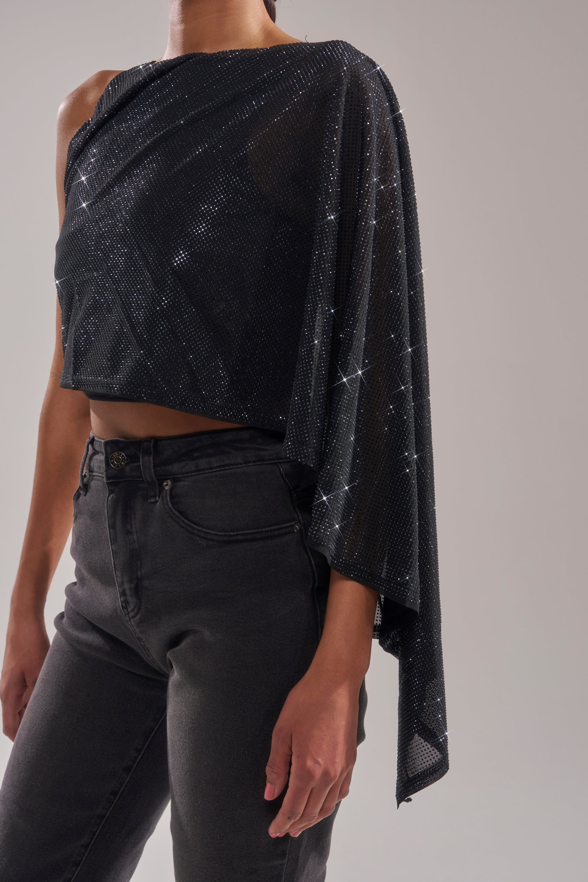 A person wearing the TOO HOT FOR YOU RHINESTONE MESH TOP—sparkly black mesh with an asymmetrical one-shoulder neckline and draped sleeve—paired with high-waisted black jeans, against a plain light background. 9