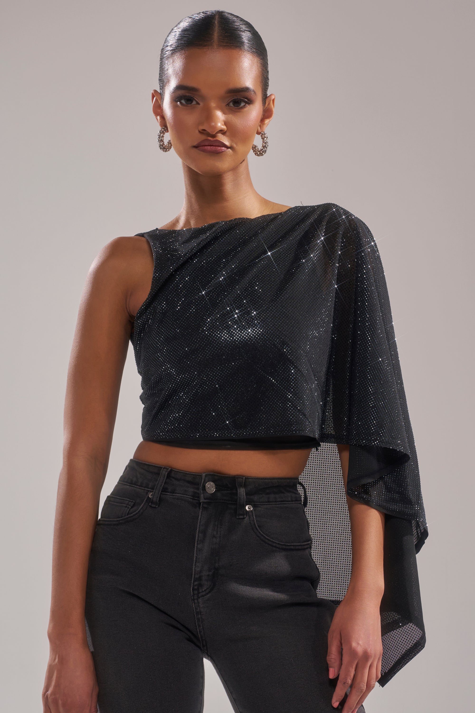 A woman with slicked-back hair models the TOO HOT FOR YOU RHINESTONE MESH TOP, featuring sparkly rhinestone mesh, a one-shoulder design with an asymmetrical draped sleeve, styled with high-waisted black jeans against a neutral background. 1