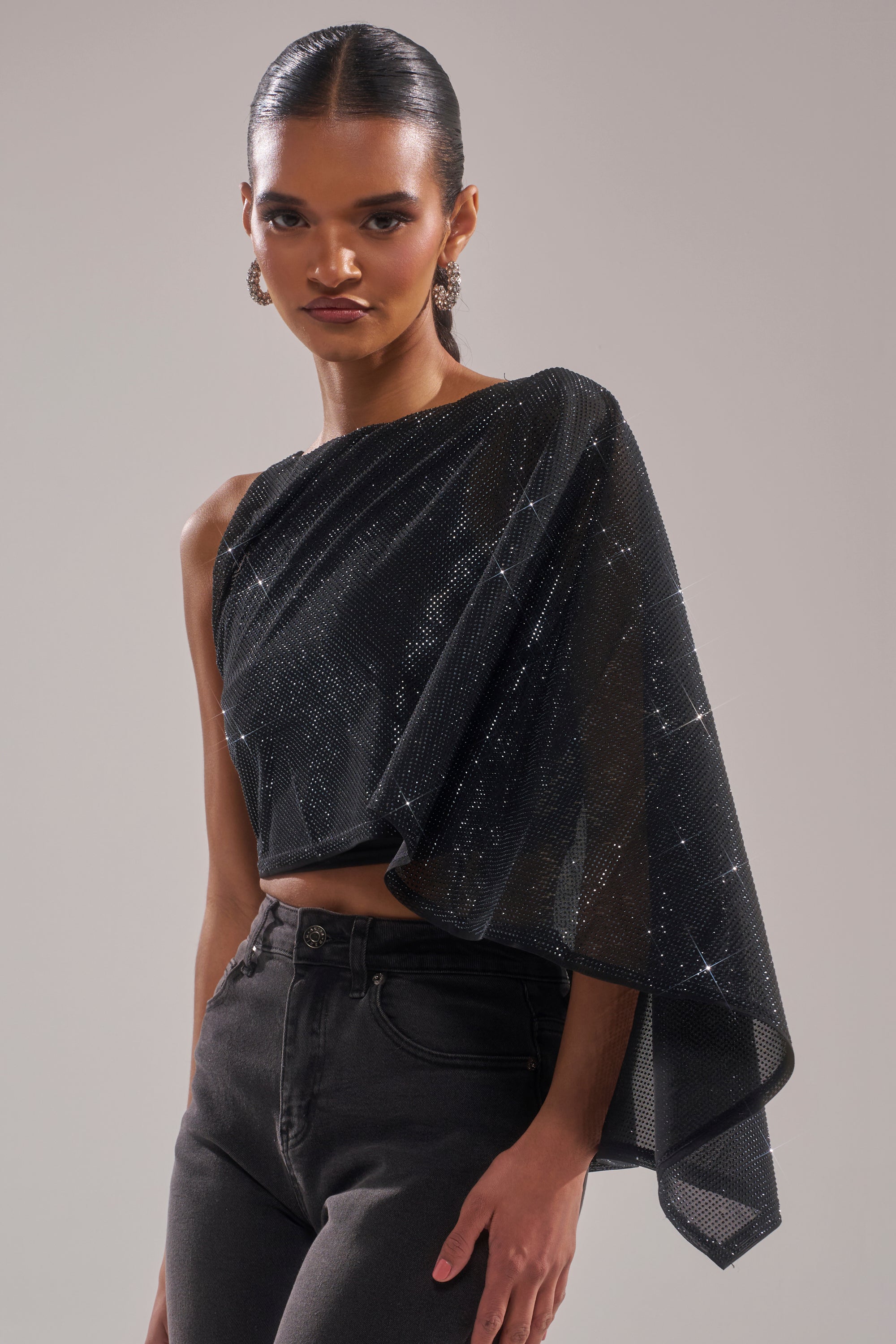 A woman poses confidently in the TOO HOT FOR YOU RHINESTONE MESH TOP, a black one-shoulder embellished style with an asymmetrical draped sleeve, paired with high-waisted black jeans and slicked-back hair. 2