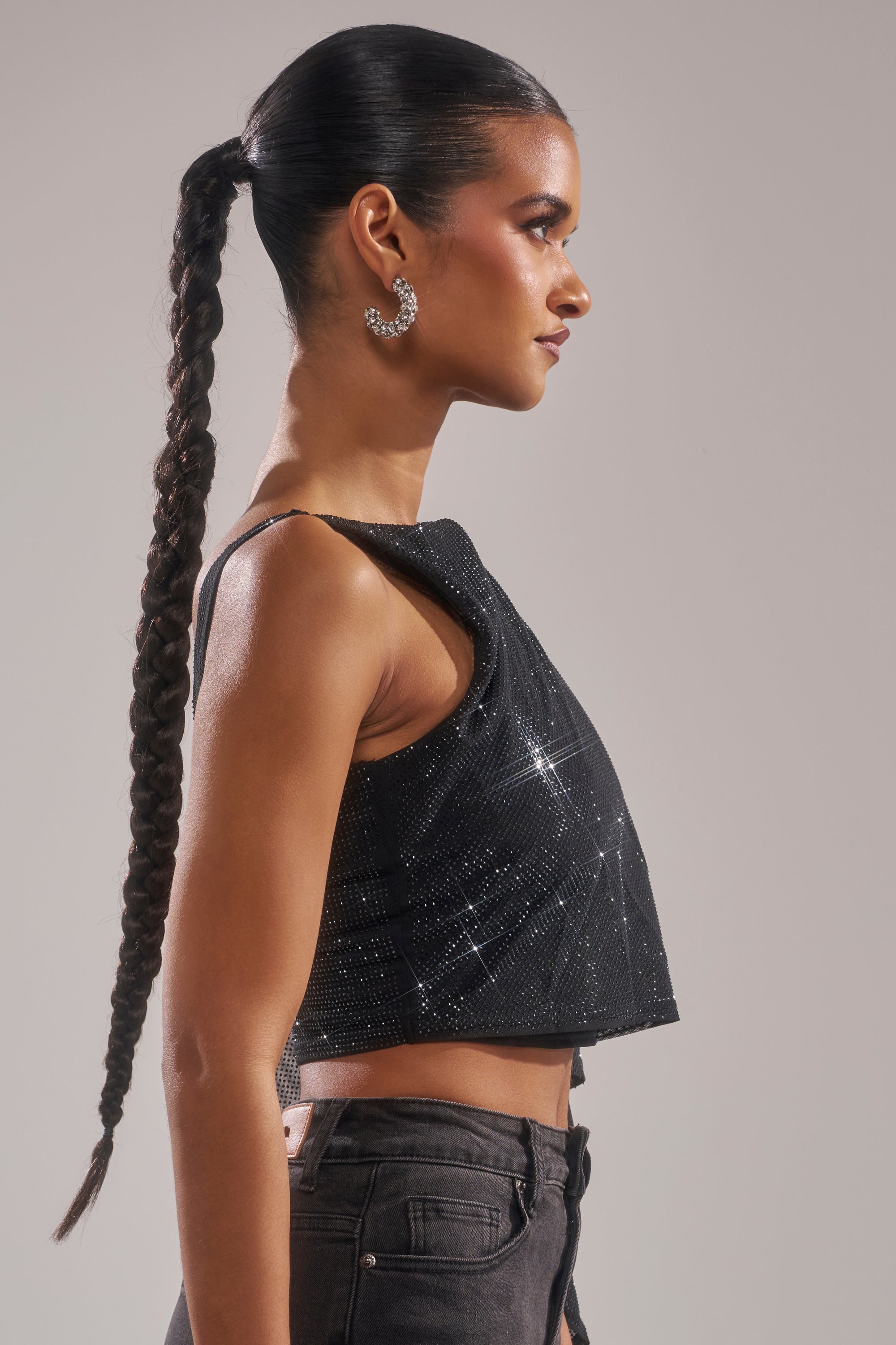 A woman with a long braided ponytail is shown in profile wearing the TOO HOT FOR YOU RHINESTONE MESH TOP, dark jeans, and large hoop earrings against a plain light background. 3