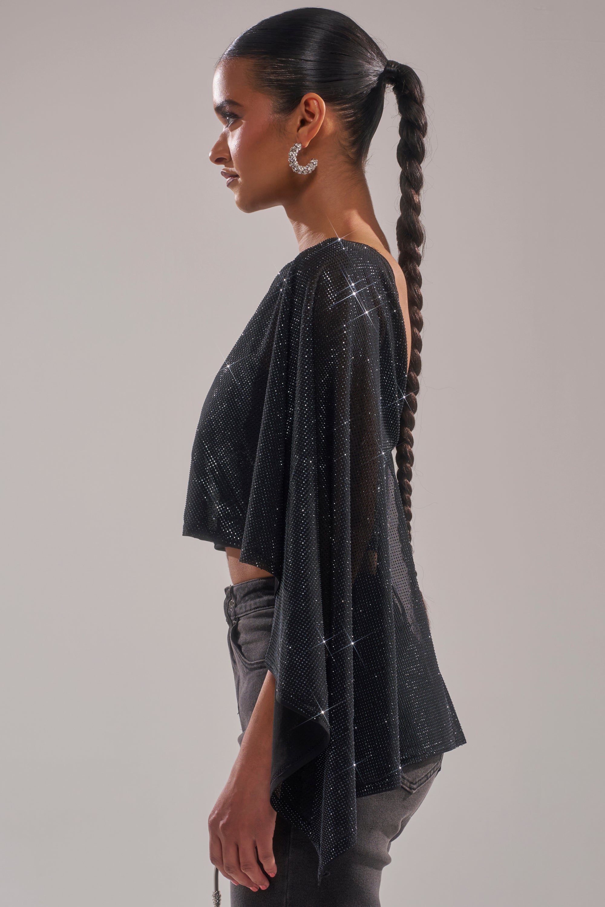 A person with a long braided ponytail wears the TOO HOT FOR YOU RHINESTONE MESH TOP featuring flowing sleeves and gray pants, standing in profile against a plain light background. 4