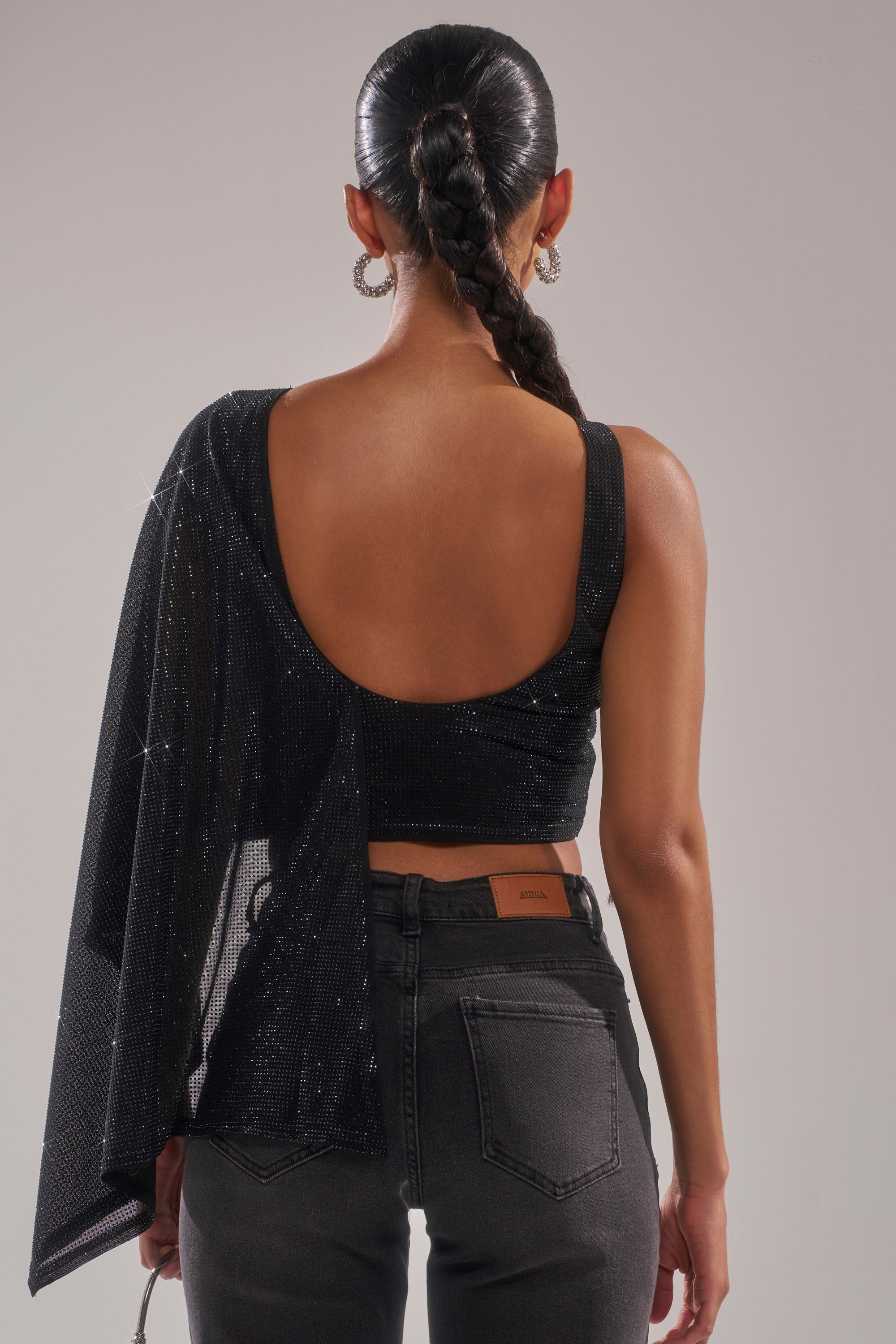 A person with a long braided ponytail is seen from the back wearing the TOO HOT FOR YOU RHINESTONE MESH TOP, featuring a one-shoulder neckline and open back, paired with high-waisted dark jeans. 5